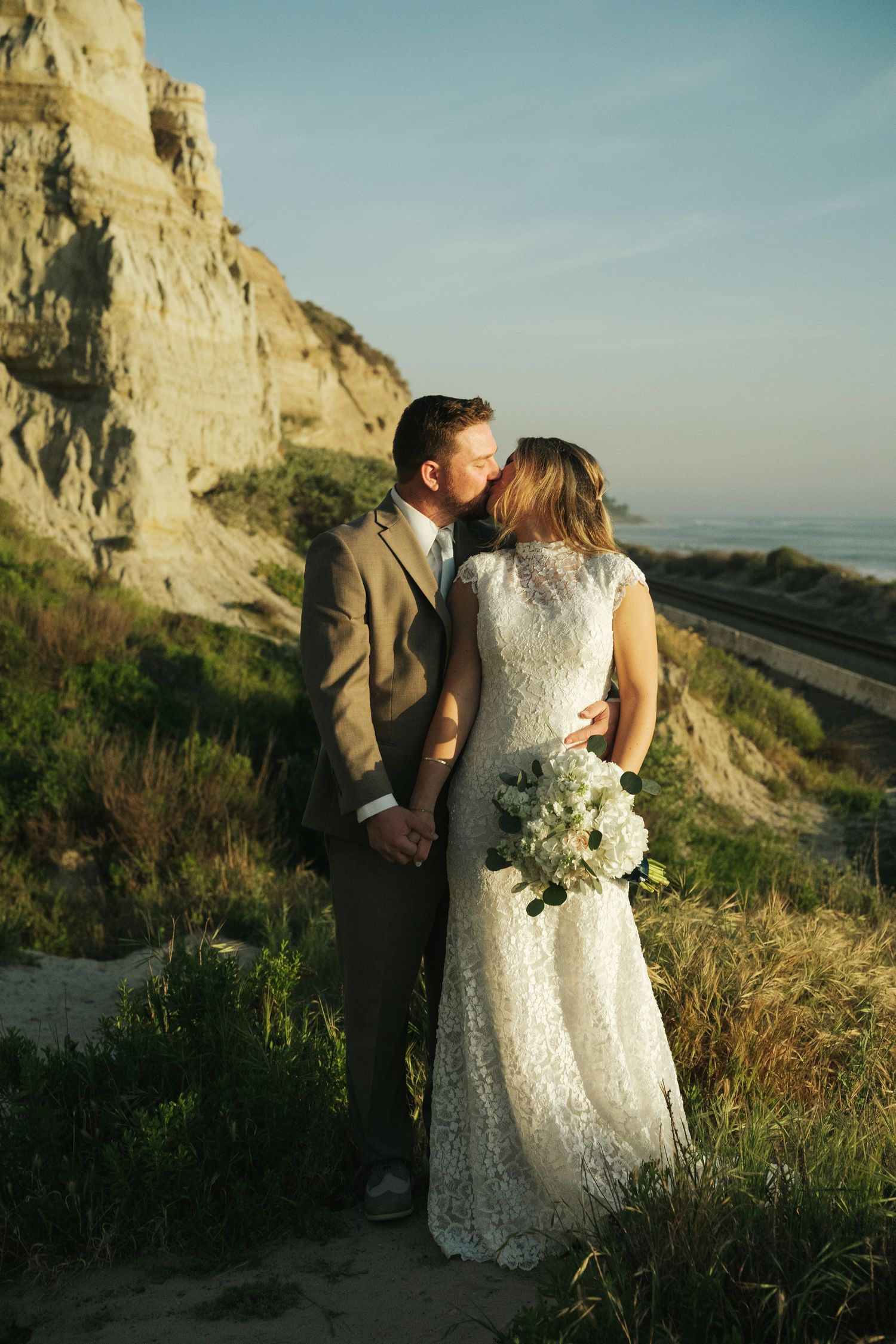 Groom kissing bride on cheek near coastal cliffs at sunset, bride holding white floral bouquet in lace gown.