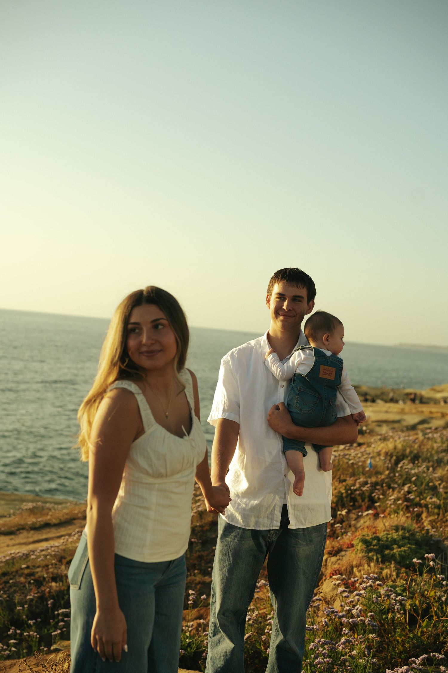 Young family with baby stands on coastal cliff at sunset, parents in jeans and white tops with ocean view behind them.