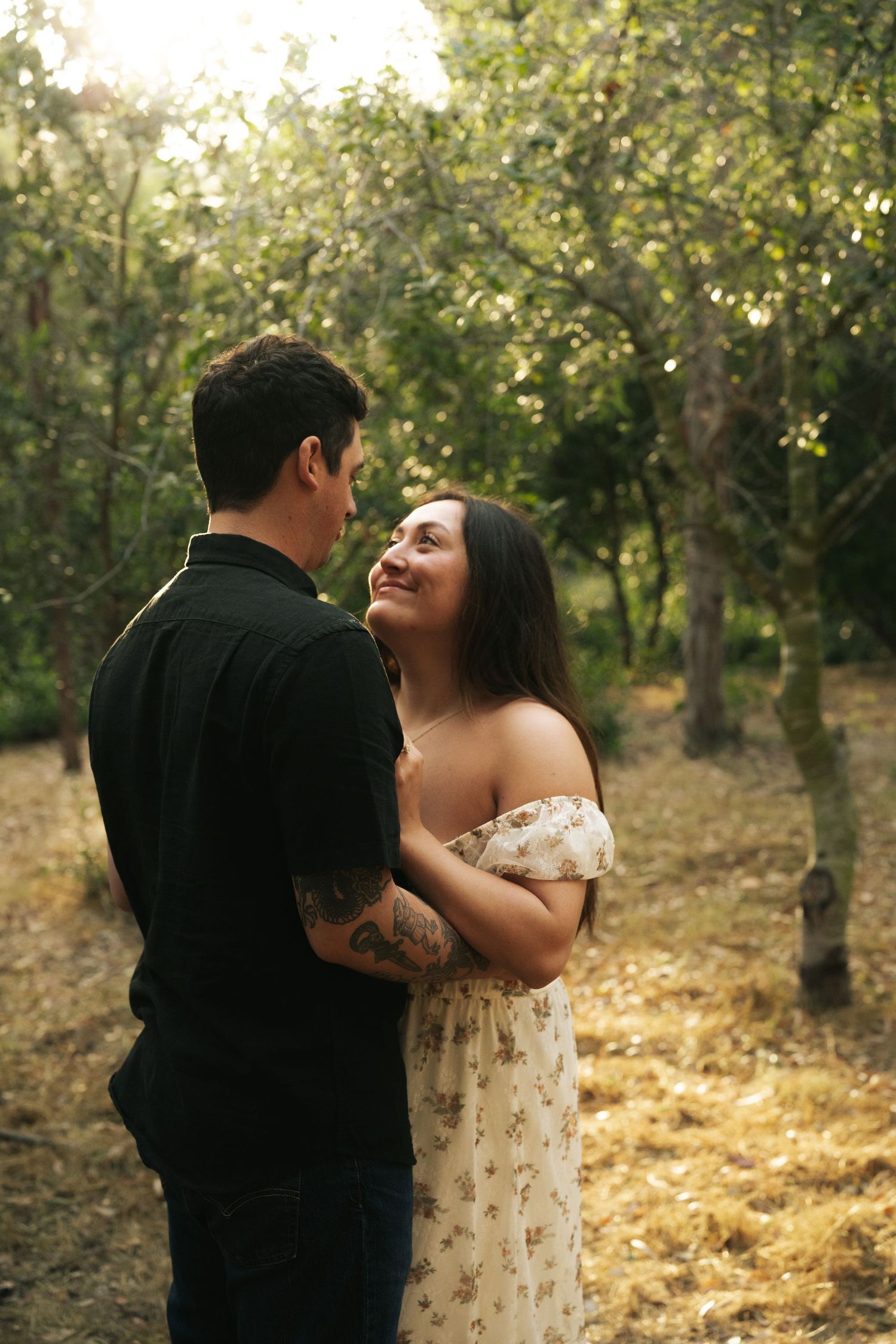 Woman in off-shoulder floral dress smiling up at partner while embracing in a sunlit forest.
