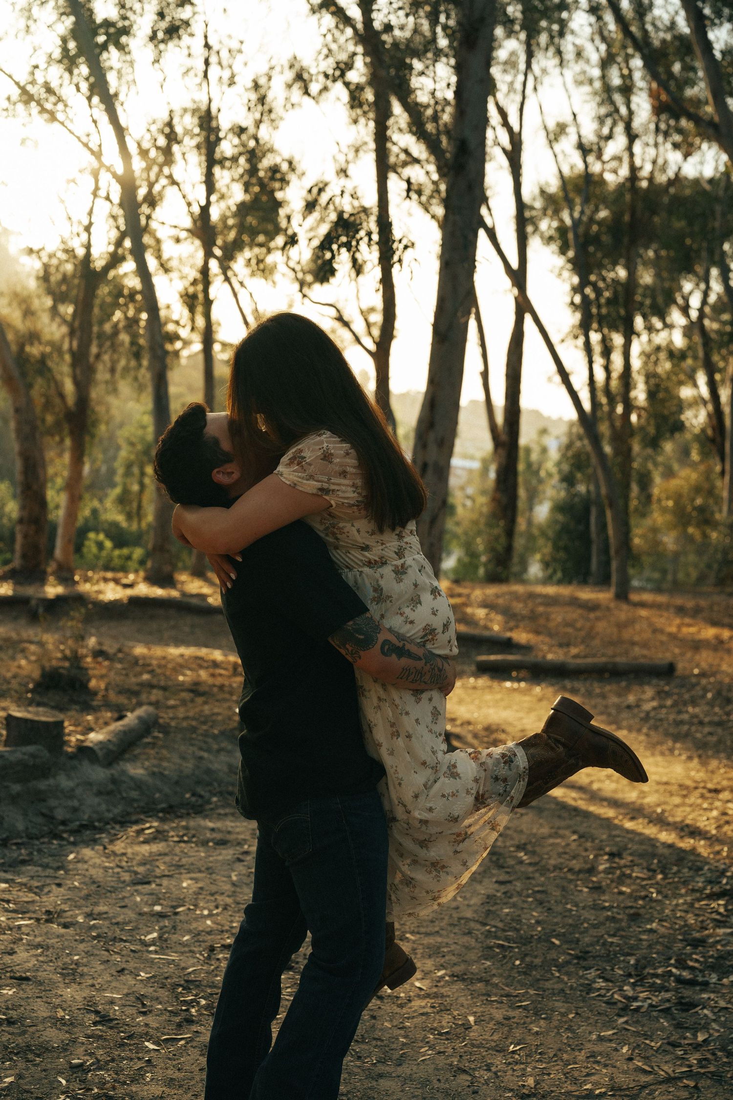 Man lifting woman in floral dress for a kiss in a sun-drenched forest at golden hour.