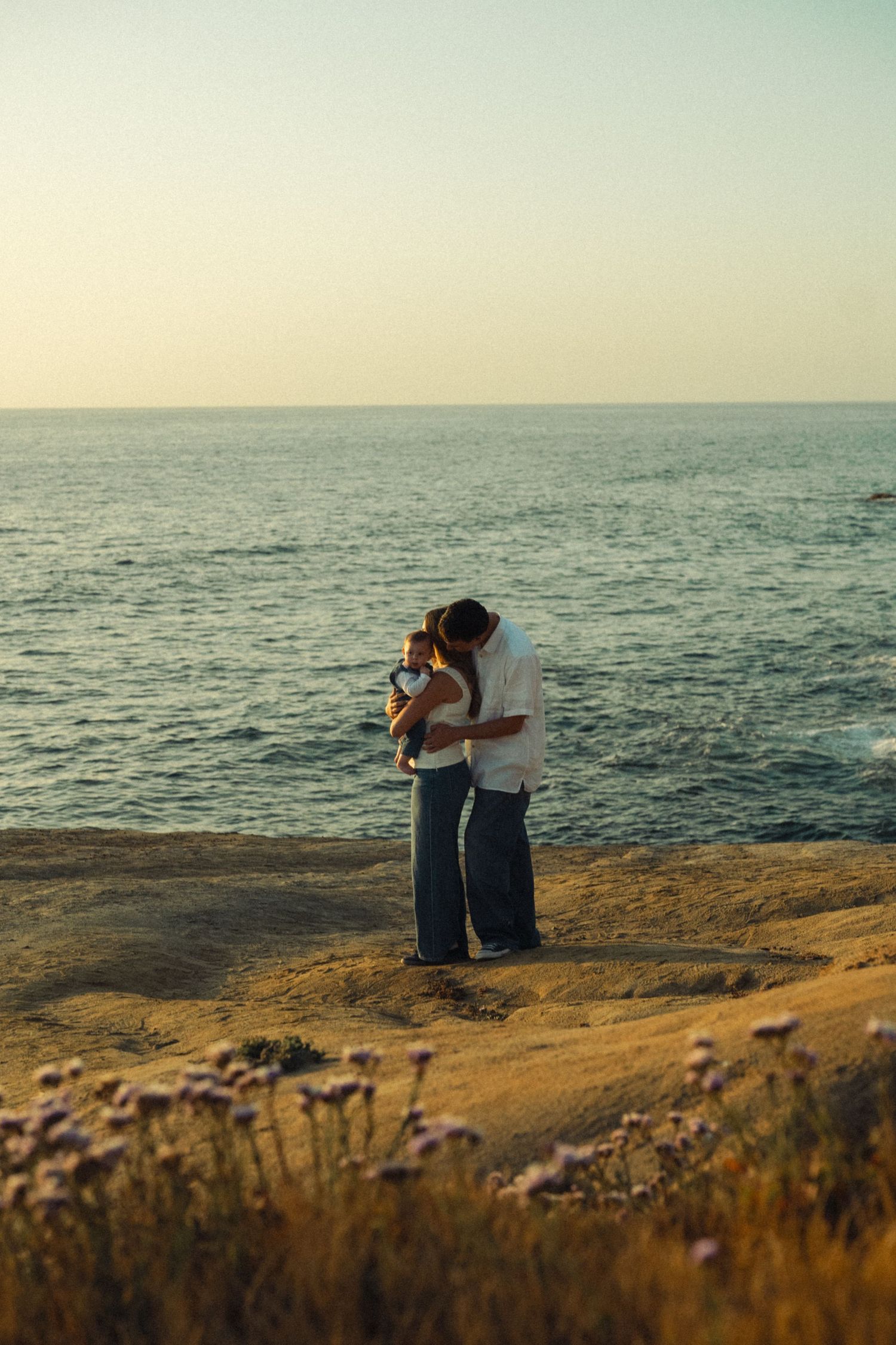 Couple embracing on a coastal cliff at sunset with wildflowers and ocean horizon in the background.