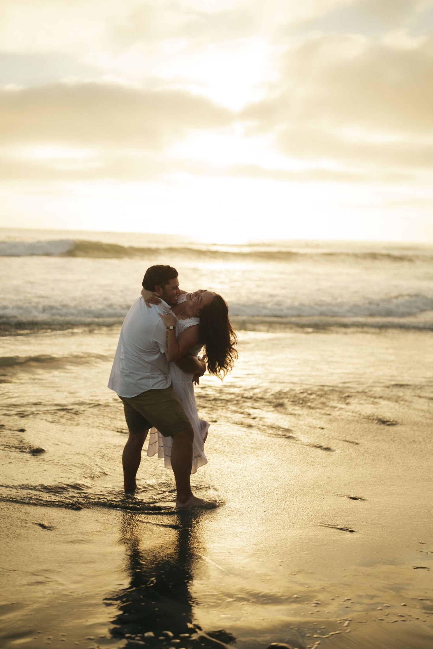 Romantic couple kissing on beach at sunset, waves reflecting golden light on wet sand behind them.