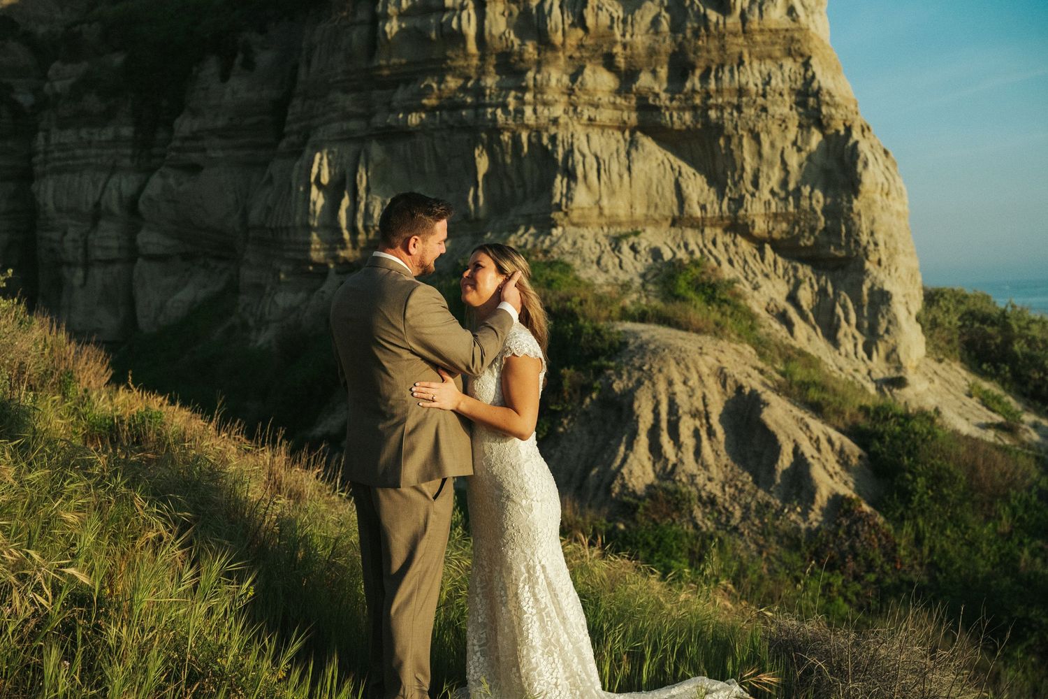 Bride and groom embrace tenderly in front of dramatic rocky cliffs on lush green hillside.