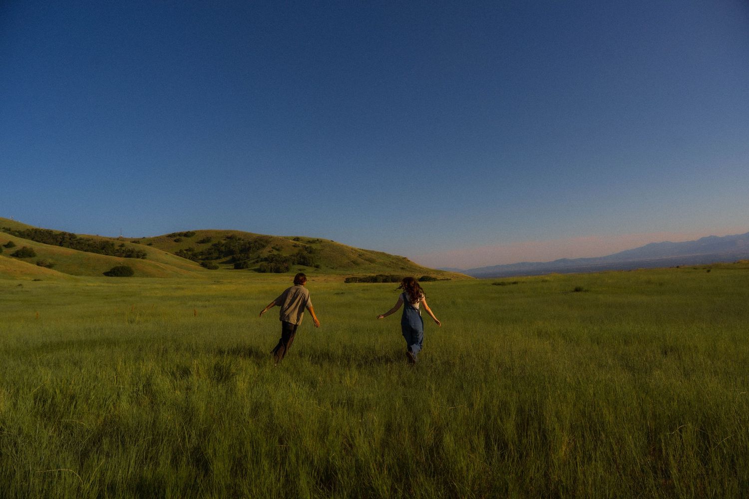 Distant figures walking through vast green meadow with rolling hills.