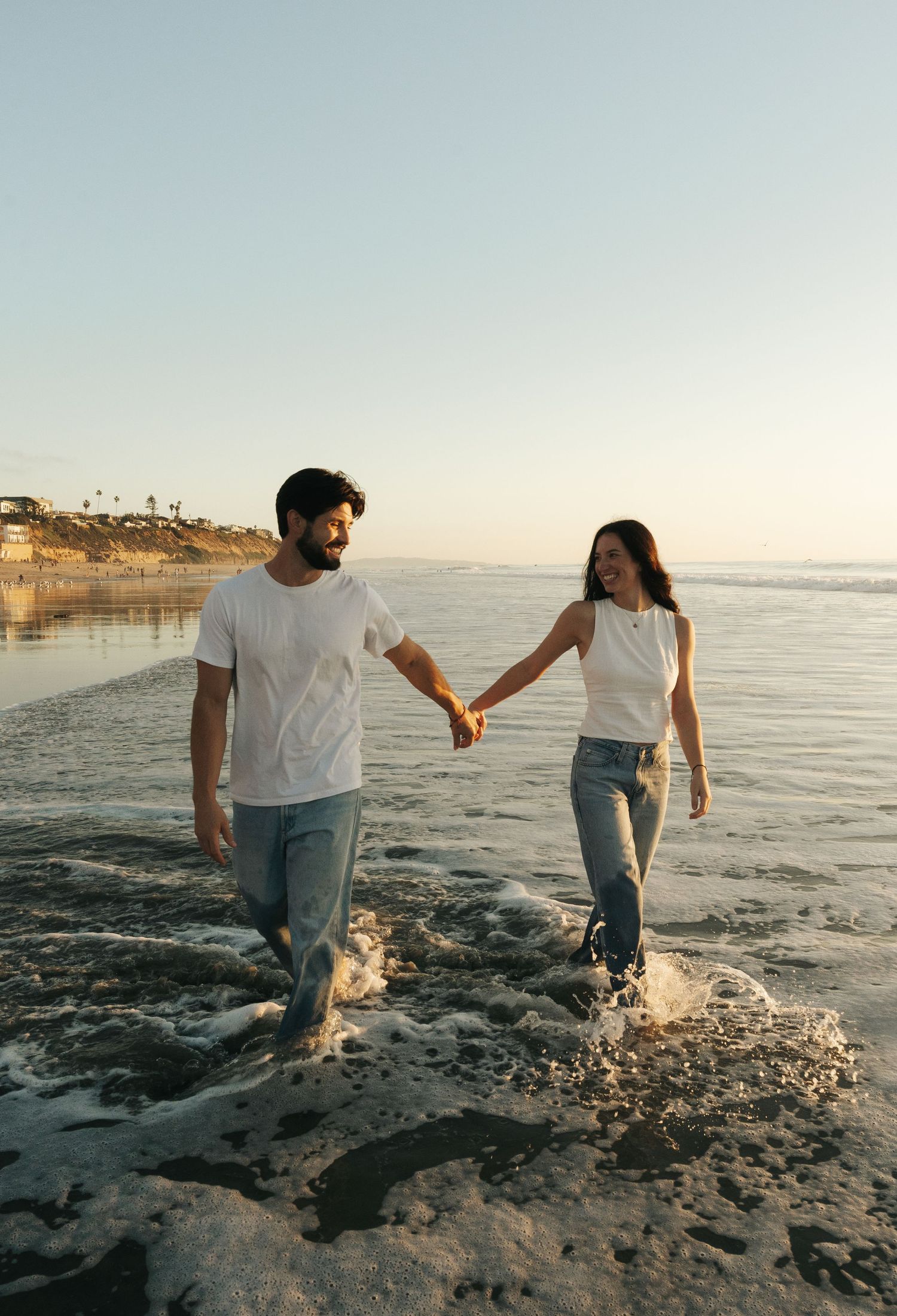 Playful couple splashing through shallow ocean water at sunset wearing matching casual outfits in Southern California.