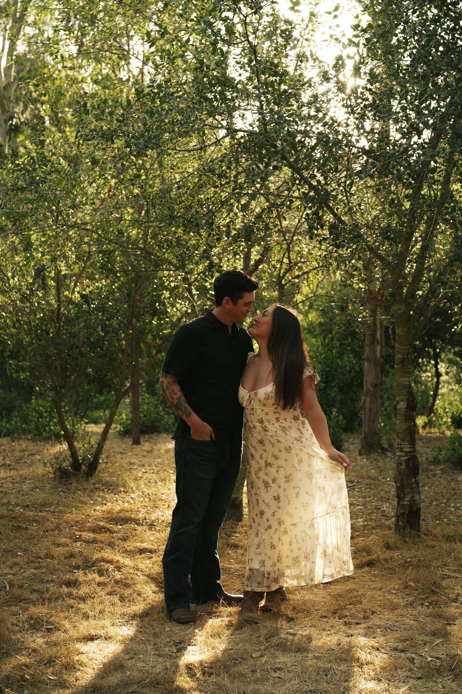 Romantic couple sharing a tender moment in golden sunlit woodland, woman in floral dress and man in black.