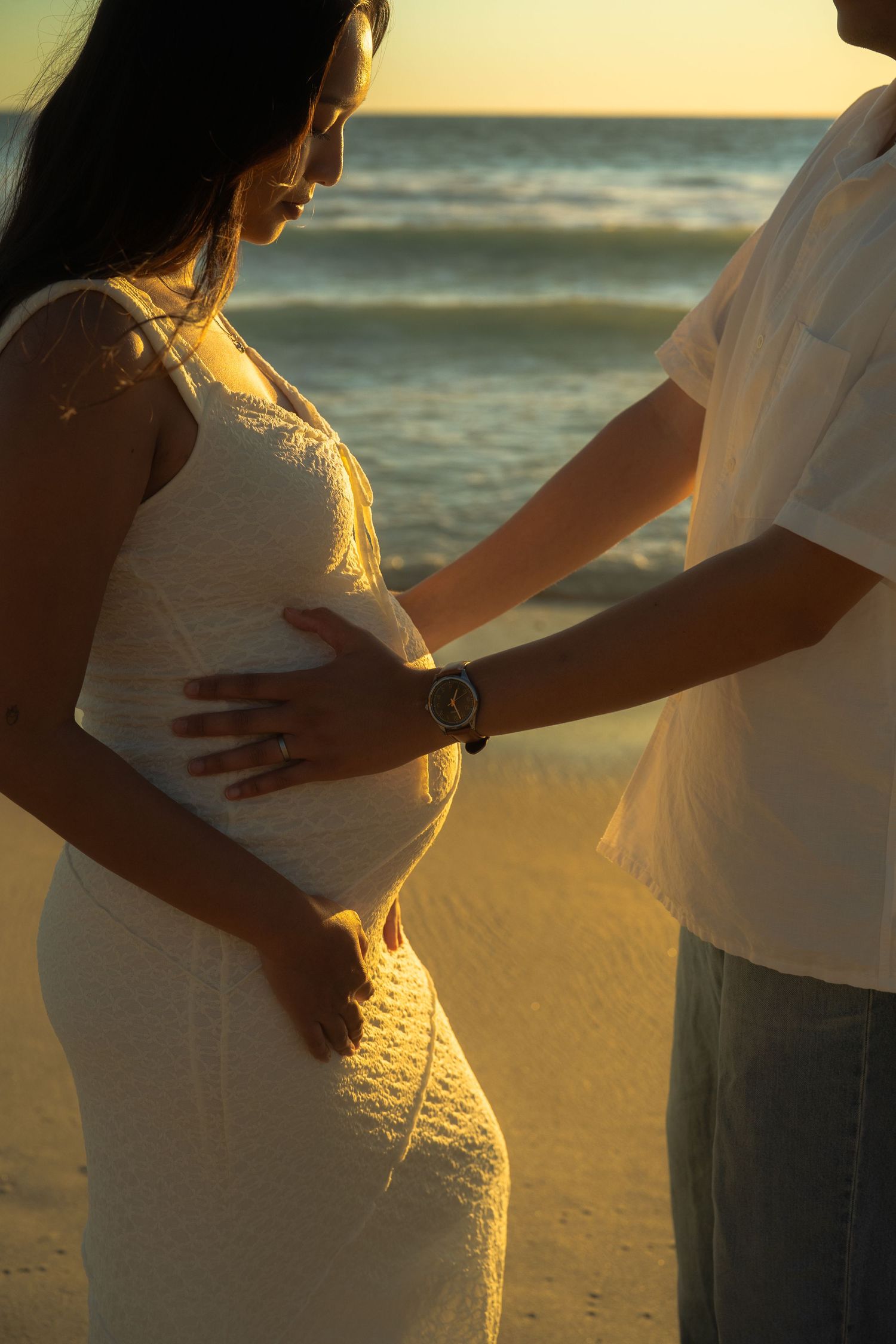 Close-up of couple tenderly holding pregnant belly at beach during warm golden sunset light.