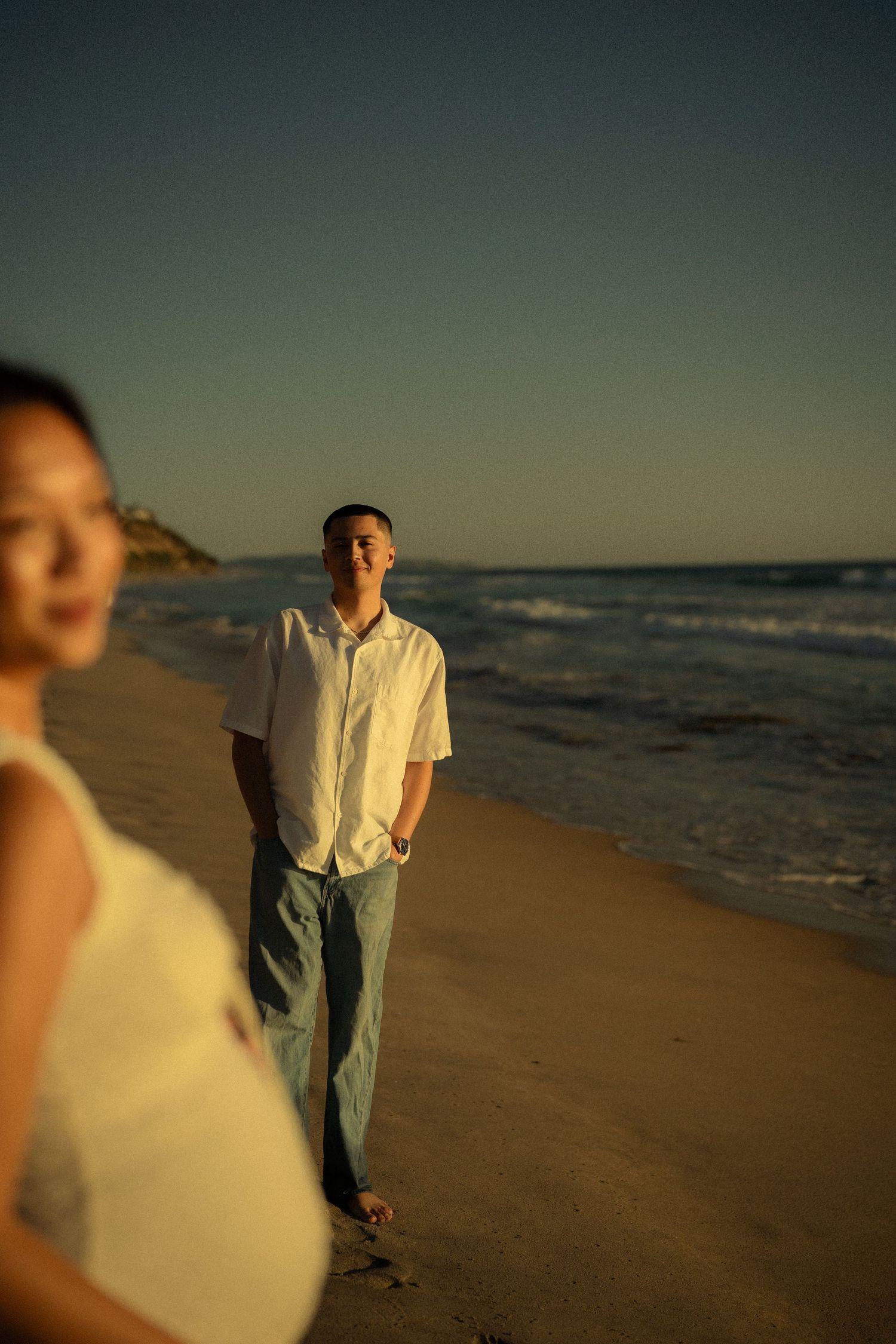 Man in white shirt walking on beach at golden hour, pregnant woman in foreground, ocean backdrop.