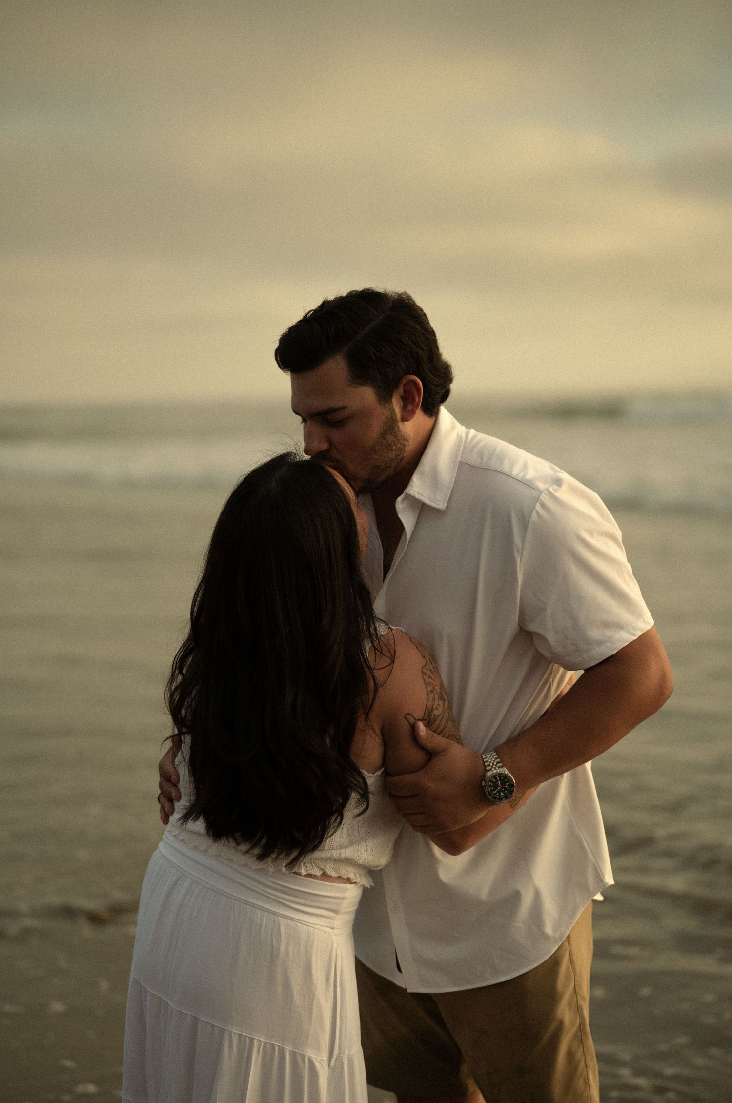 Couple sharing a romantic kiss on the beach at sunset, dressed in white, with ocean waves in the background.