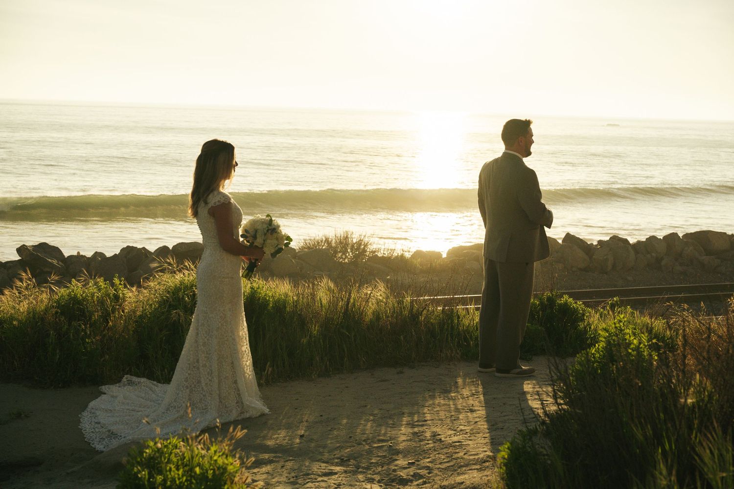 Bride and groom stand apart at sunset by the ocean, bride holding bouquet in lace gown, groom in dark suit.