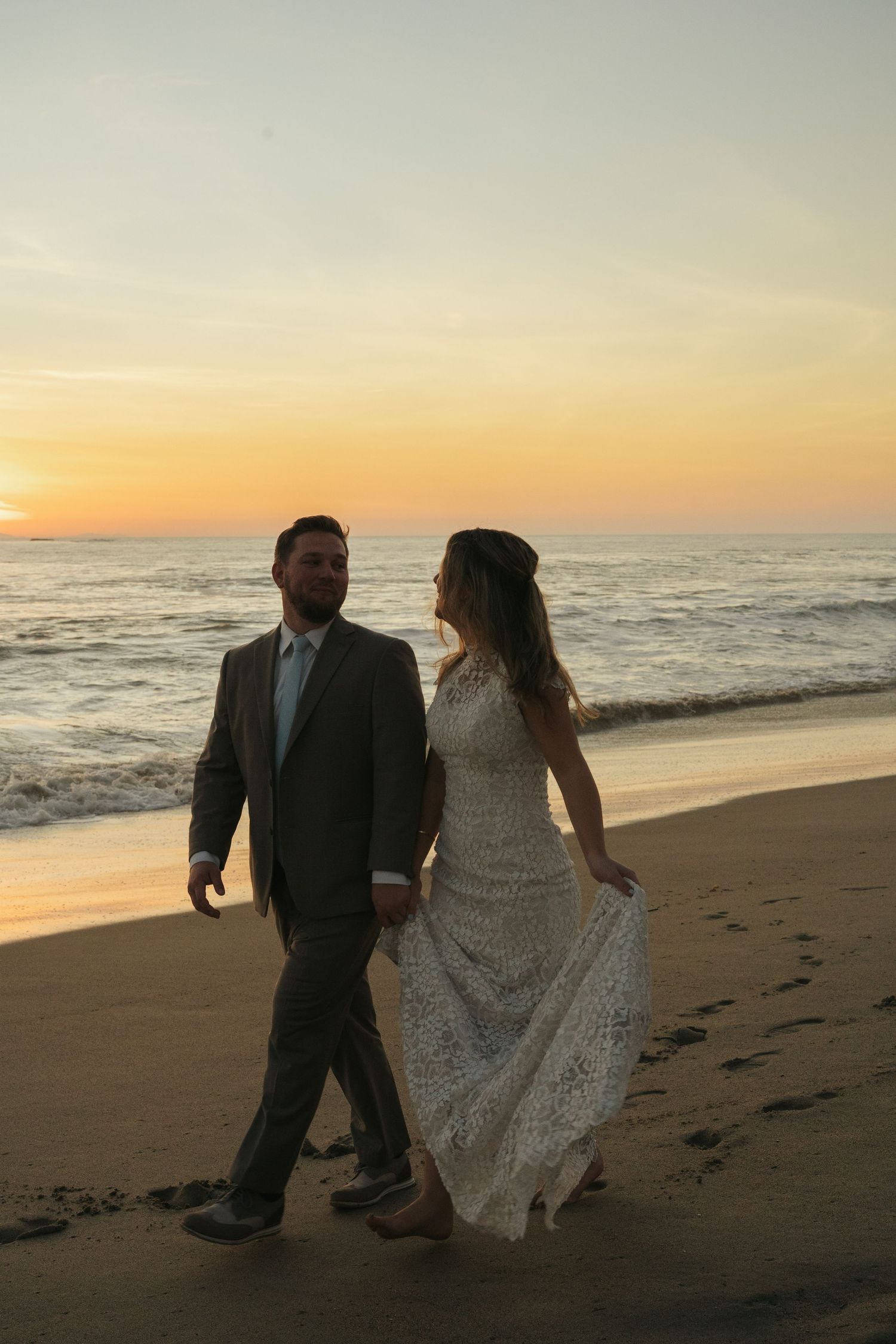 Bride and groom walk hand-in-hand along sandy beach at sunset, bride in lace wedding gown.