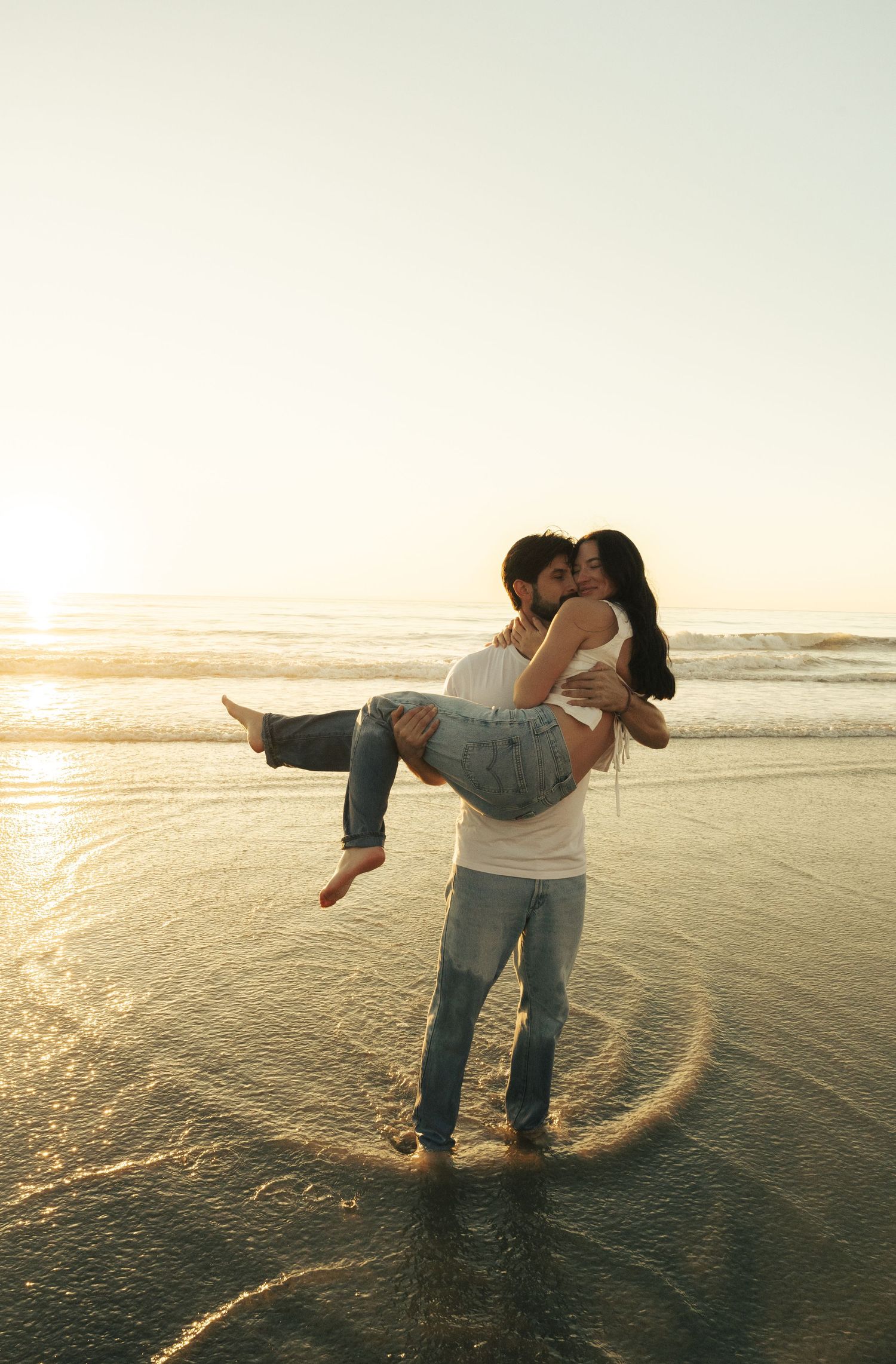 Couple embracing on a beach at sunset with golden light reflecting off wet sand and ocean waves.