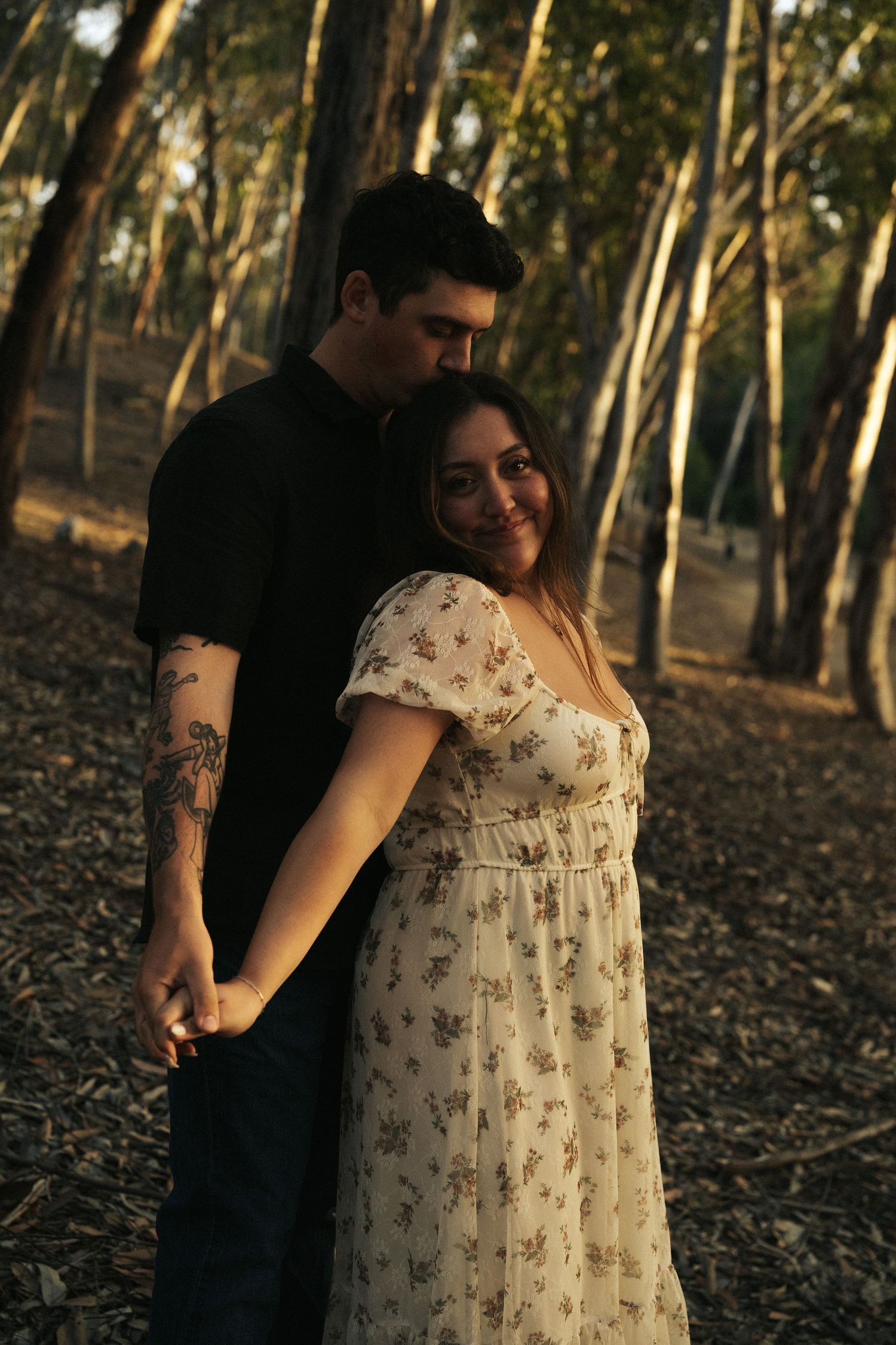 Man in black shirt embracing woman in floral dress from behind in a sun-dappled forest, she smiles at camera.