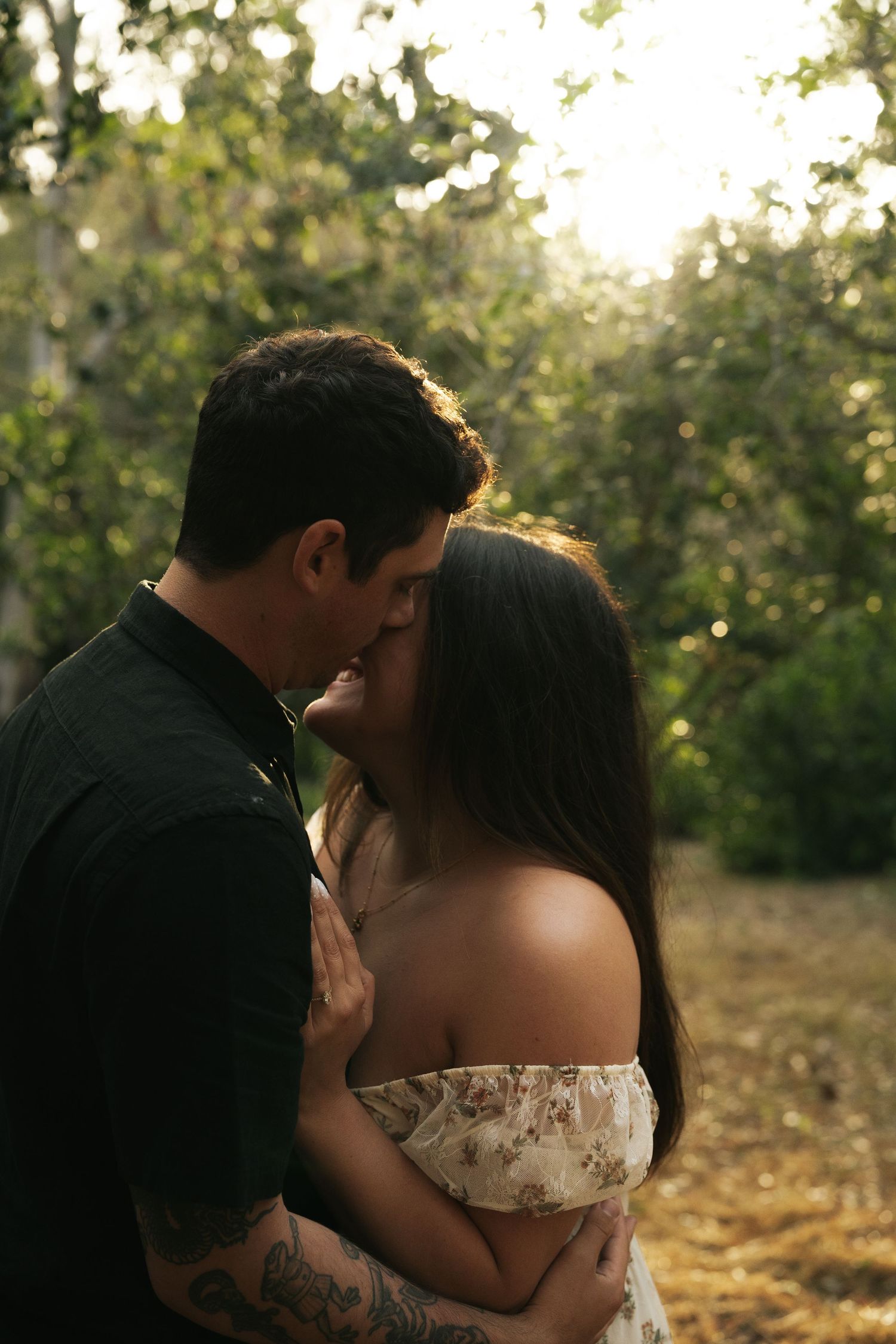 An engaged couple shares a romantic kiss outdoors, bathed in warm golden sunlight filtering through green trees.
