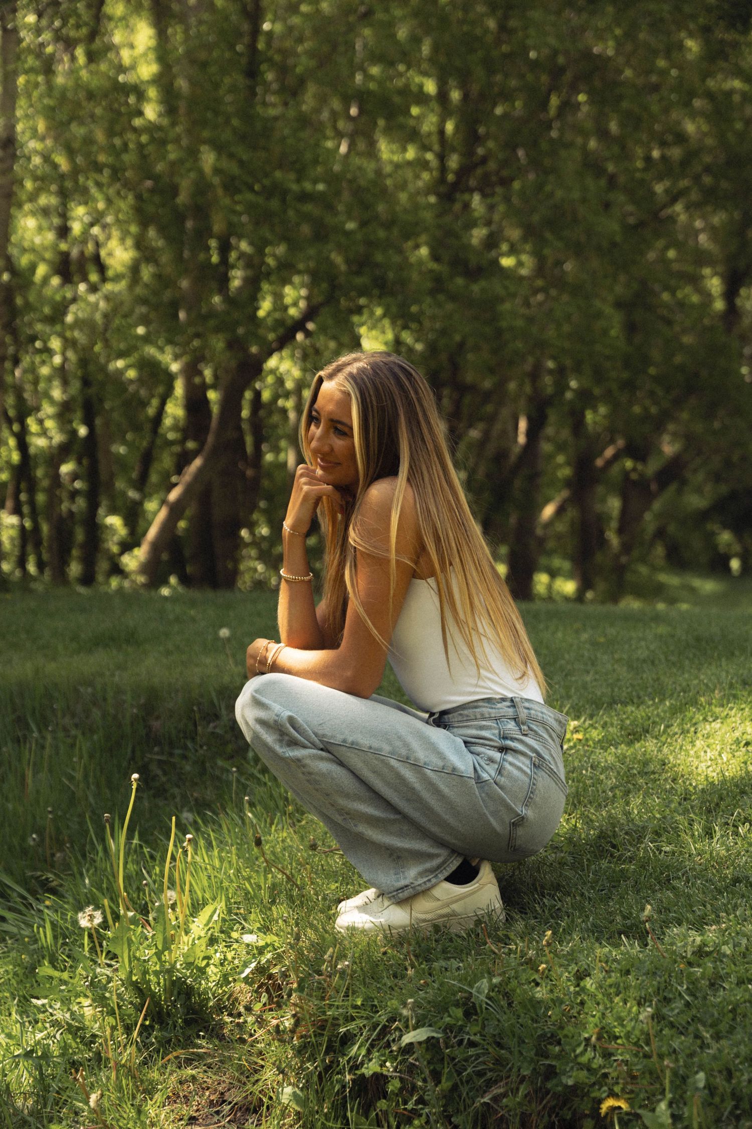 A casual portrait of someone crouching in a sunlit grassy area wearing jeans and a white top.