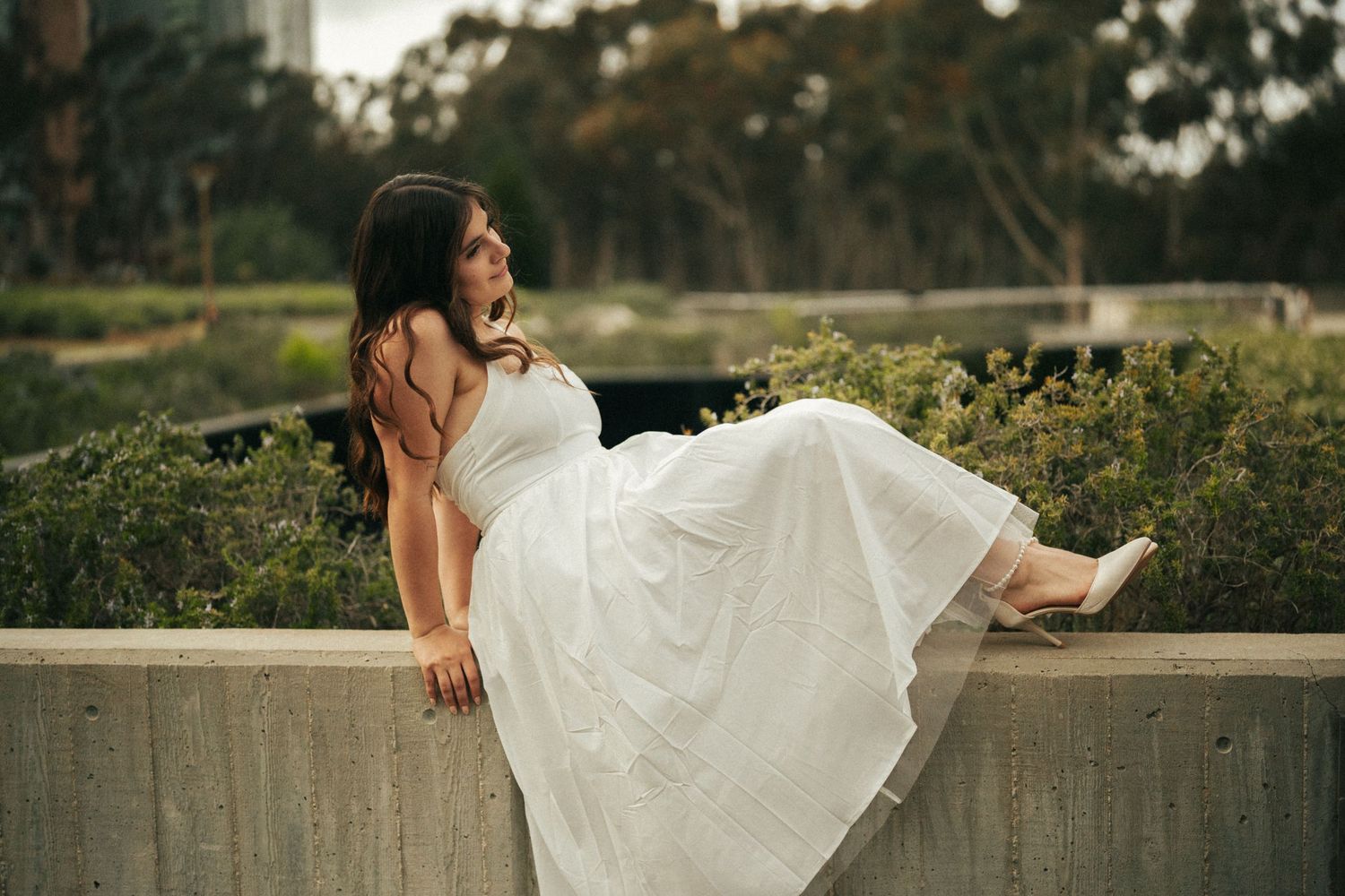 Woman in white dress sitting on concrete wall surrounded by lush greenery in an outdoor setting.