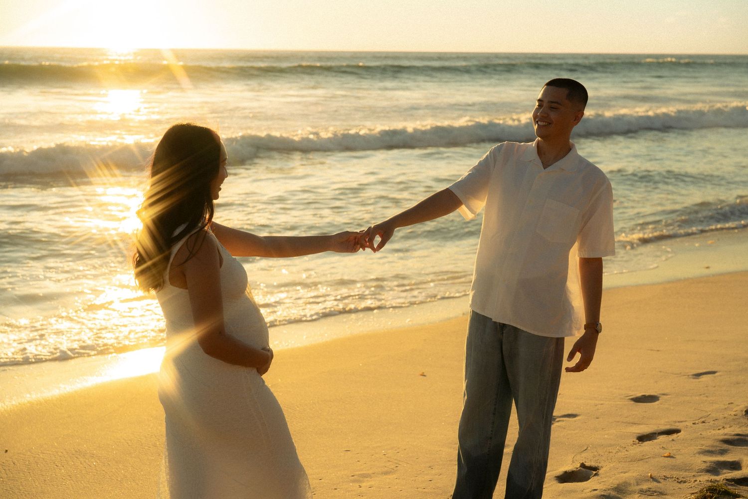 Pregnant couple holding hands on a sunny beach at golden hour with ocean waves.