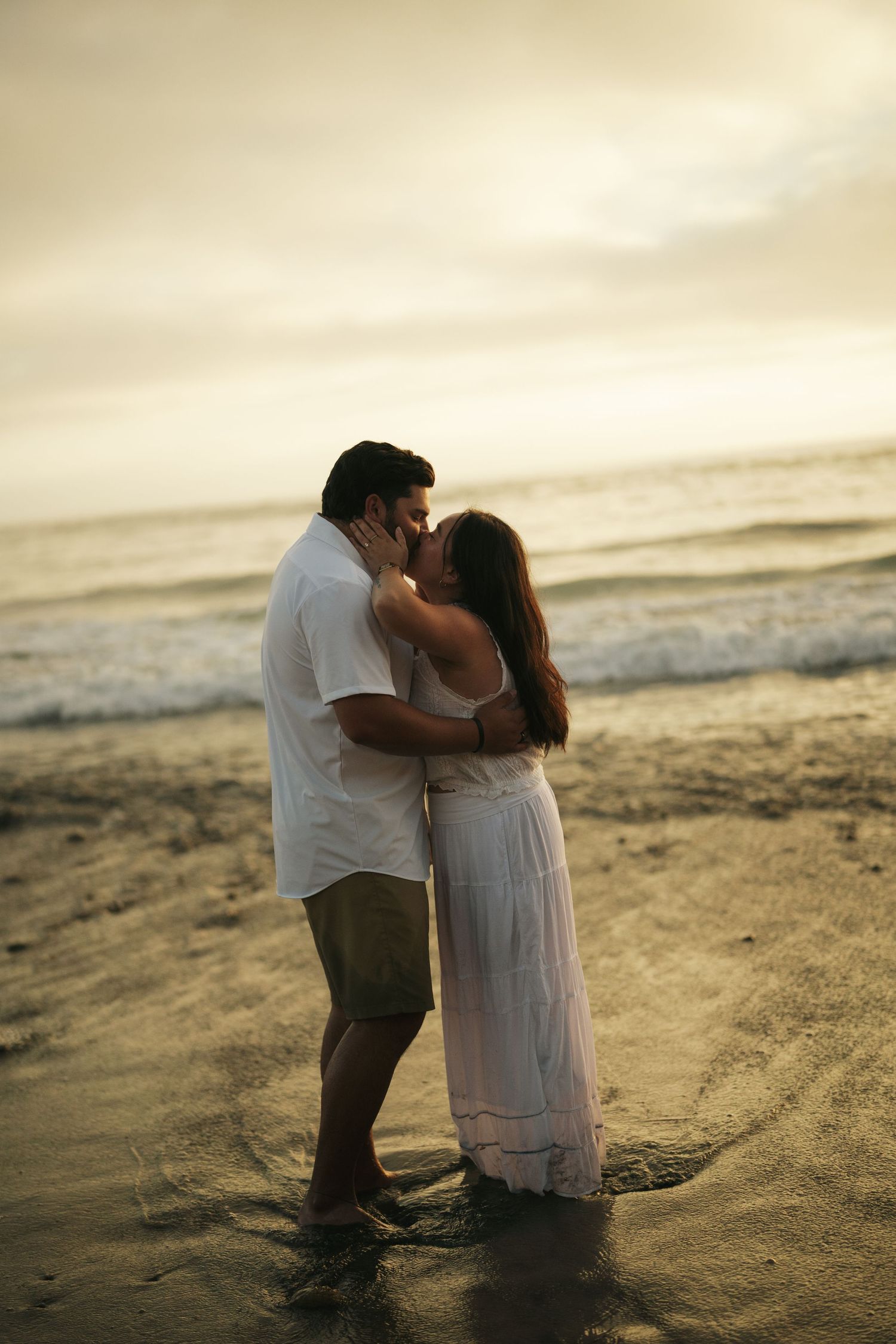 Romantic couple kissing on sandy beach at sunset, woman in flowing white dress and man in white shirt and khaki shorts.