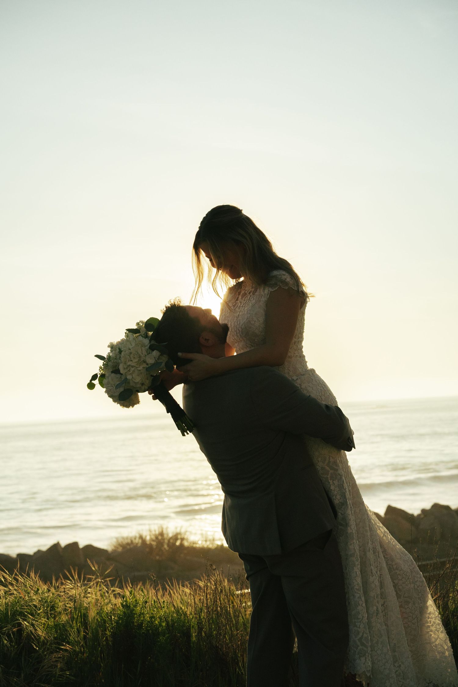 Groom lifts bride in silhouette against glowing ocean sunset, sharing romantic moment with white bouquet.