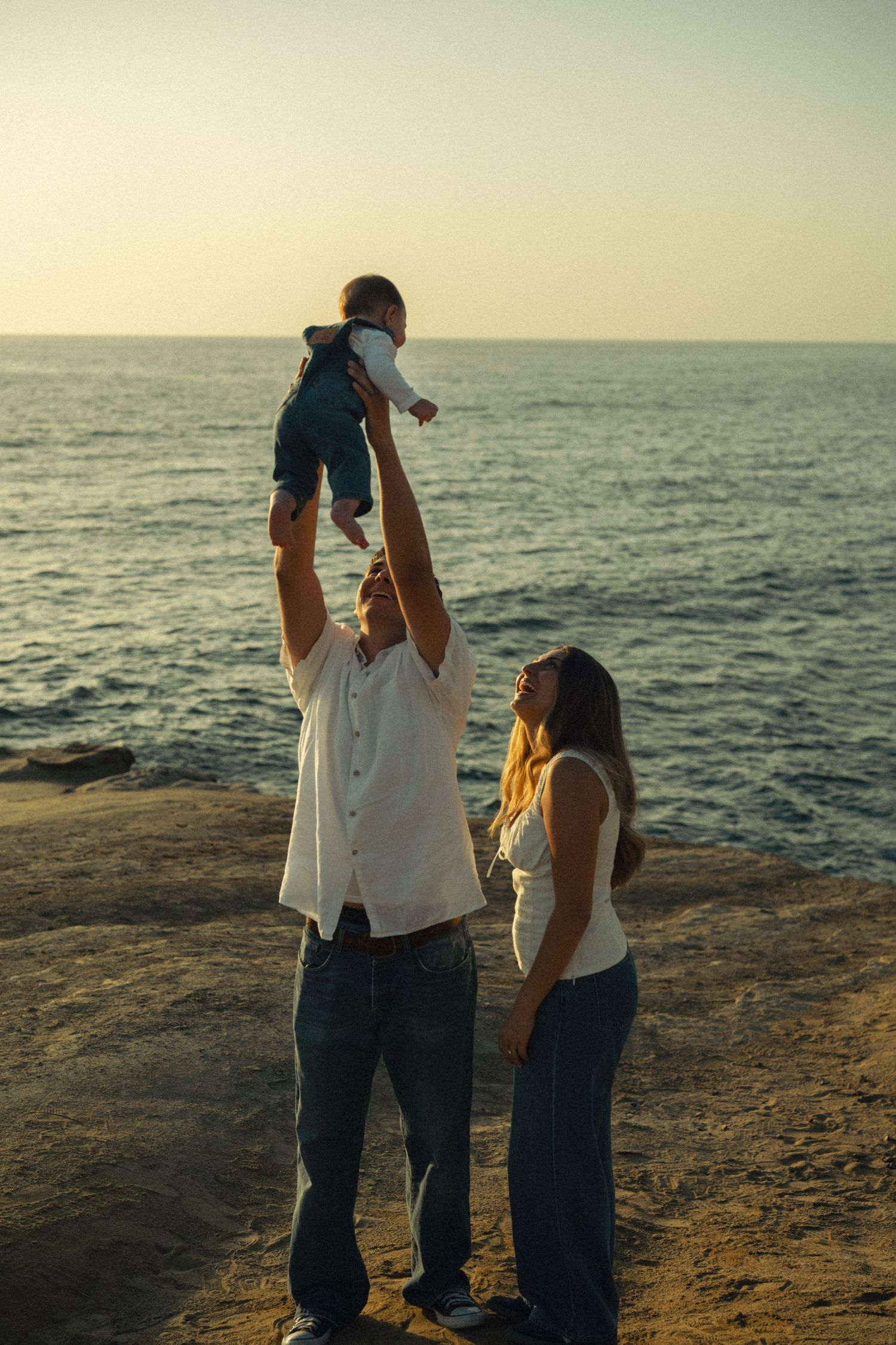 Joyful father lifts baby high in the air while mother watches by the ocean at sunset.