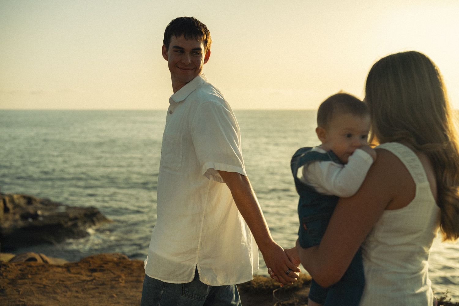 Smiling man looks back at camera while holding hands with woman carrying baby at ocean sunset.