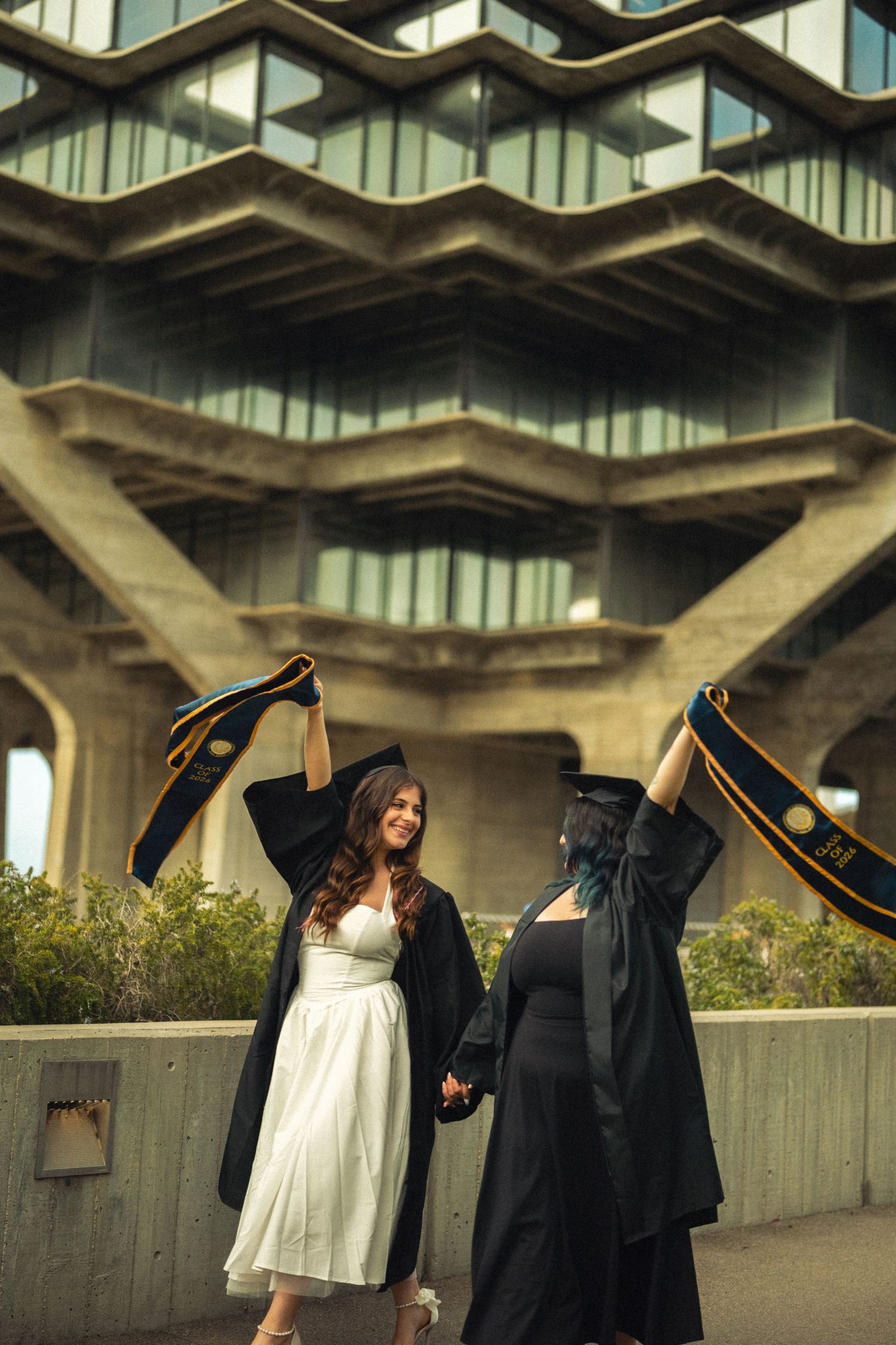 Two graduates in black gowns and caps celebrate outdoors in front of the iconic Geisel Library at UC San Diego.