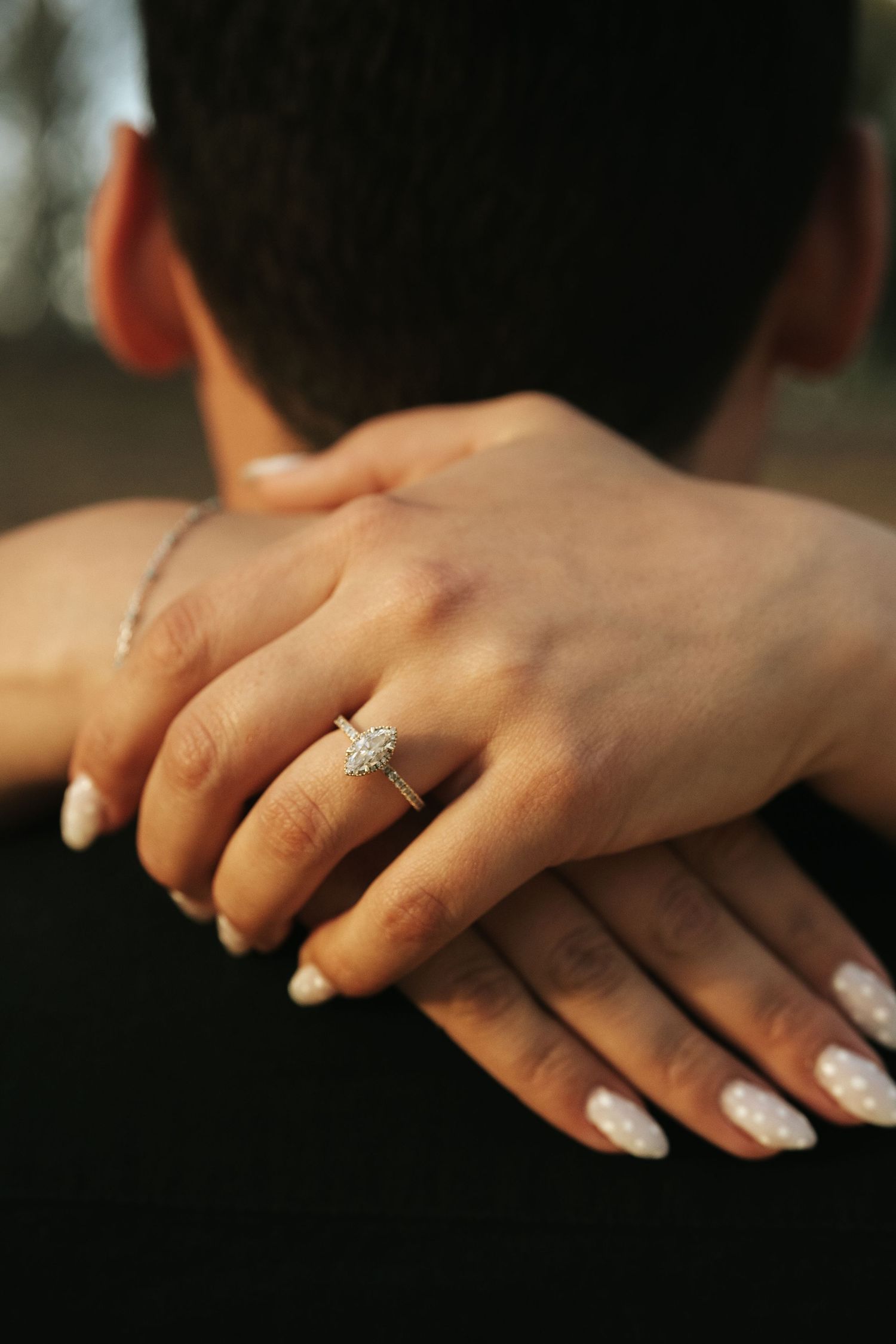 Close-up of a womans hand displaying a sparkling oval diamond engagement ring resting on a mans shoulder.