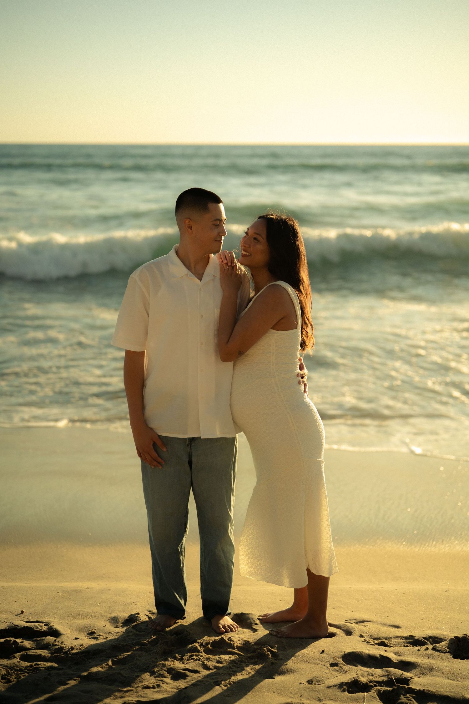 Smiling pregnant couple in white outfits standing together on sandy beach at sunset with ocean waves behind.