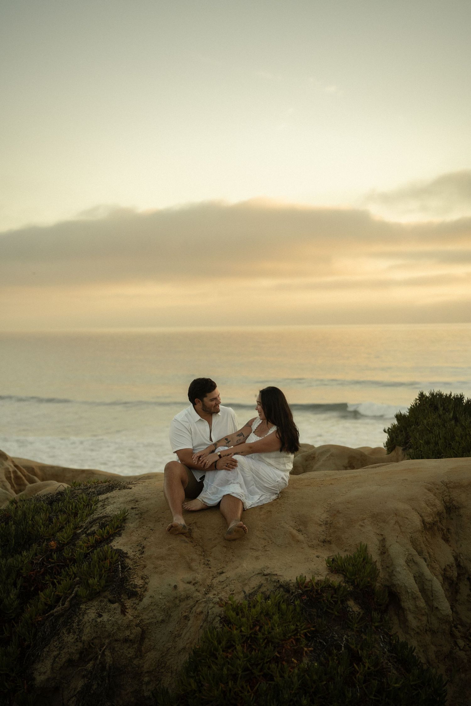Couple sitting on coastal rocks at sunset, sharing a romantic moment with ocean waves in the background.