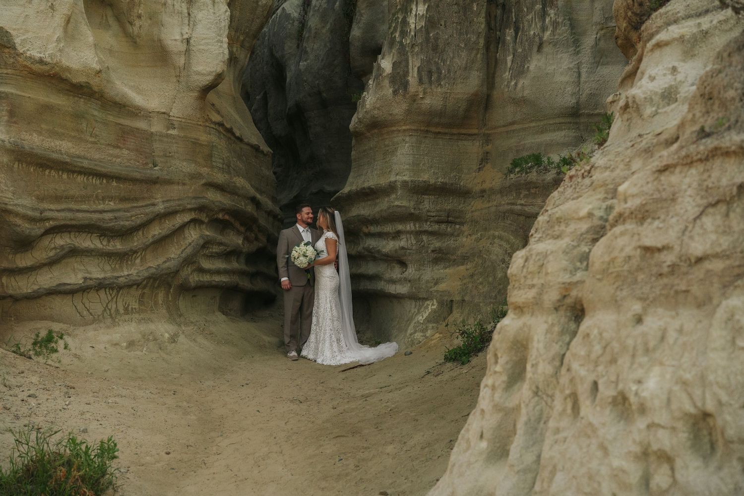 Bride and groom stand together in a stunning sandstone canyon arch, bride holding a white bouquet on their wedding day.