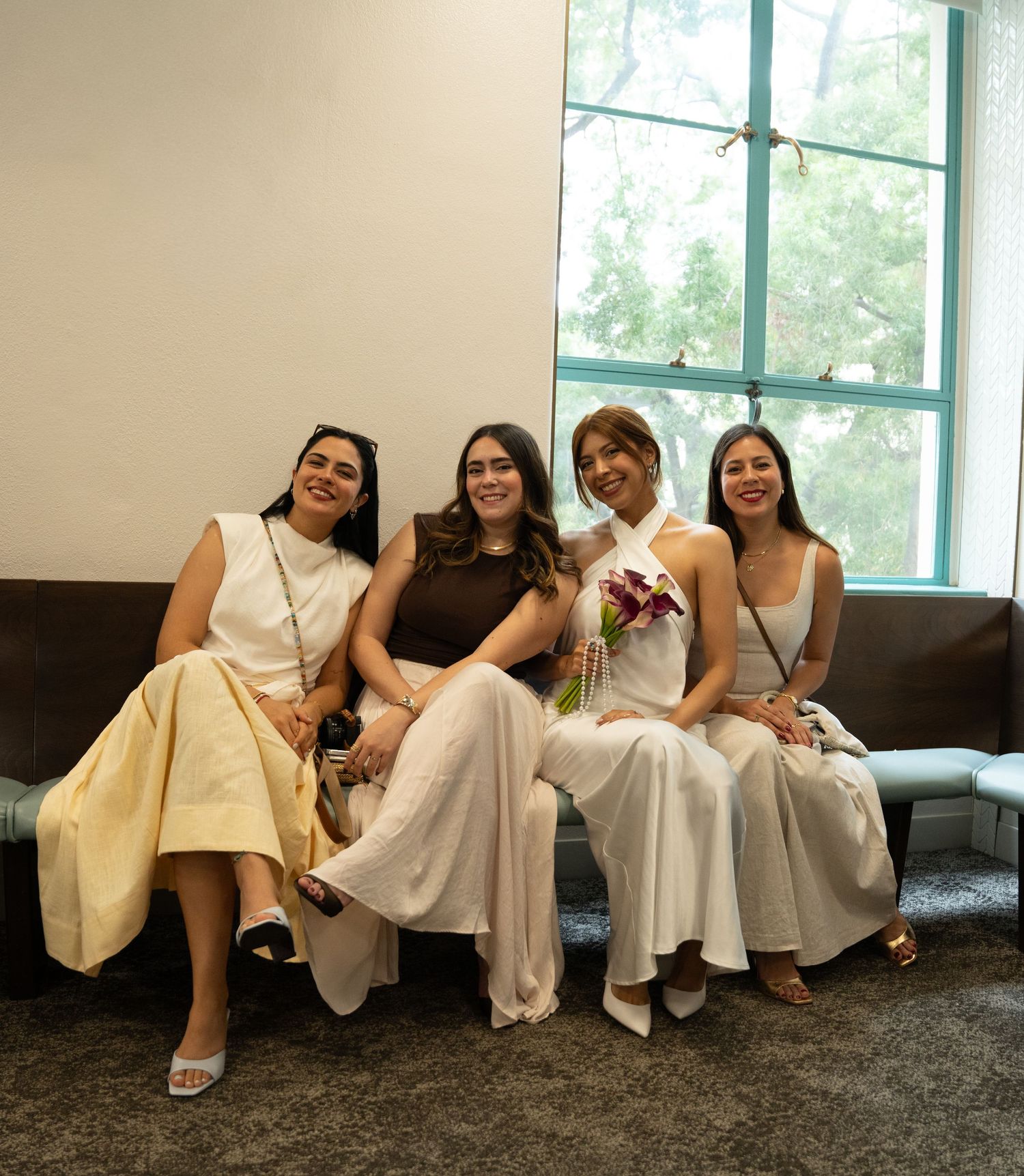 Four women in elegant white and neutral outfits smile together on a couch near a bright window at a formal event.