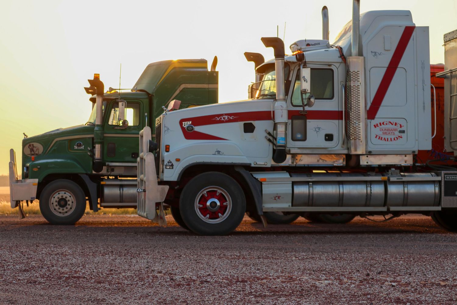Row of semi trucks and farm equipment parked at sunset against an orange sky.