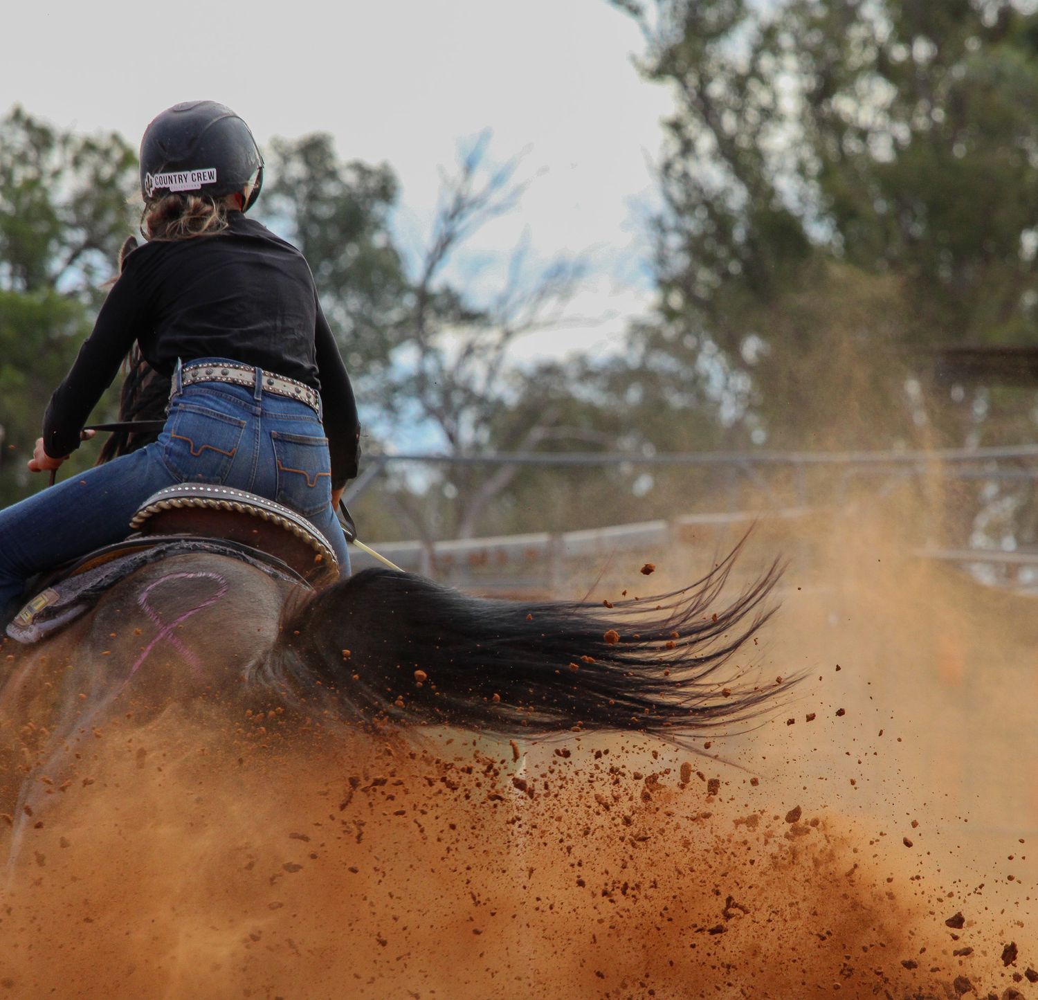 Horse and rider kick up dirt while racing at high speed during barrel racing competition in outdoor arena.
