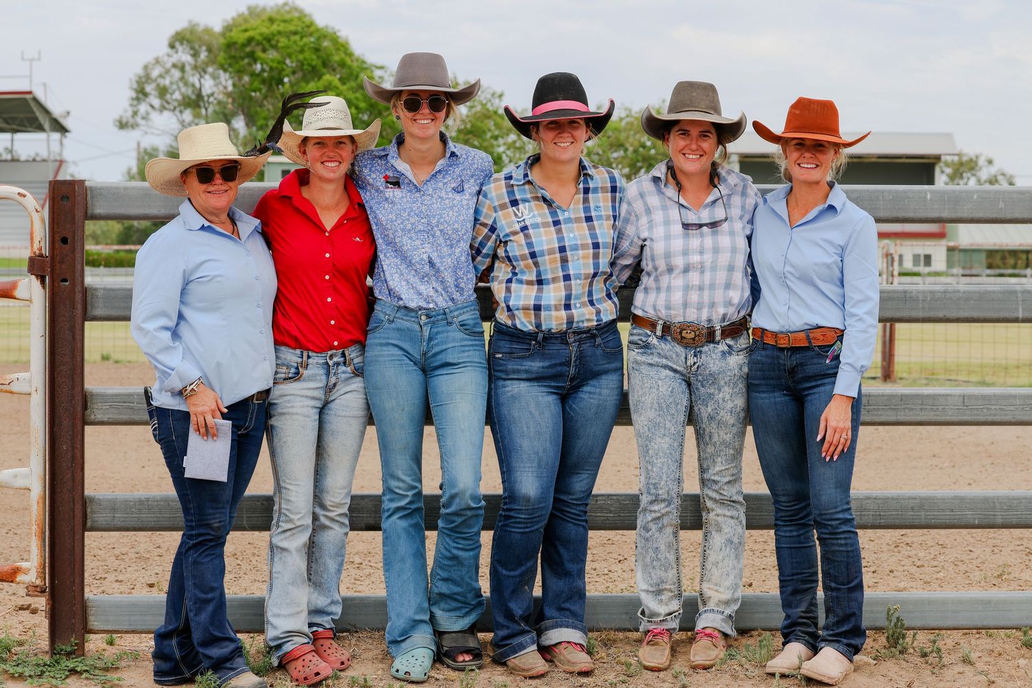 Row of people in western wear sitting casually on wooden fences at a ranch.