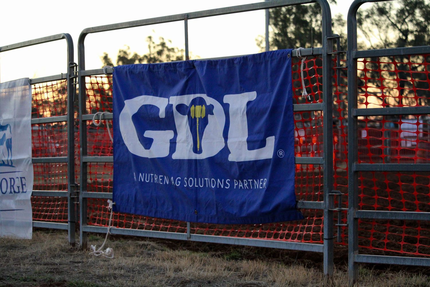 Blue banners and advertising signage hang along metal barriers at an outdoor sporting event venue at sunset.