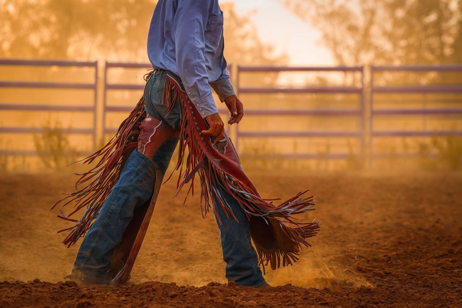 A cowboy in chaps and boots kicks up dust in a golden-lit rodeo arena at sunset.