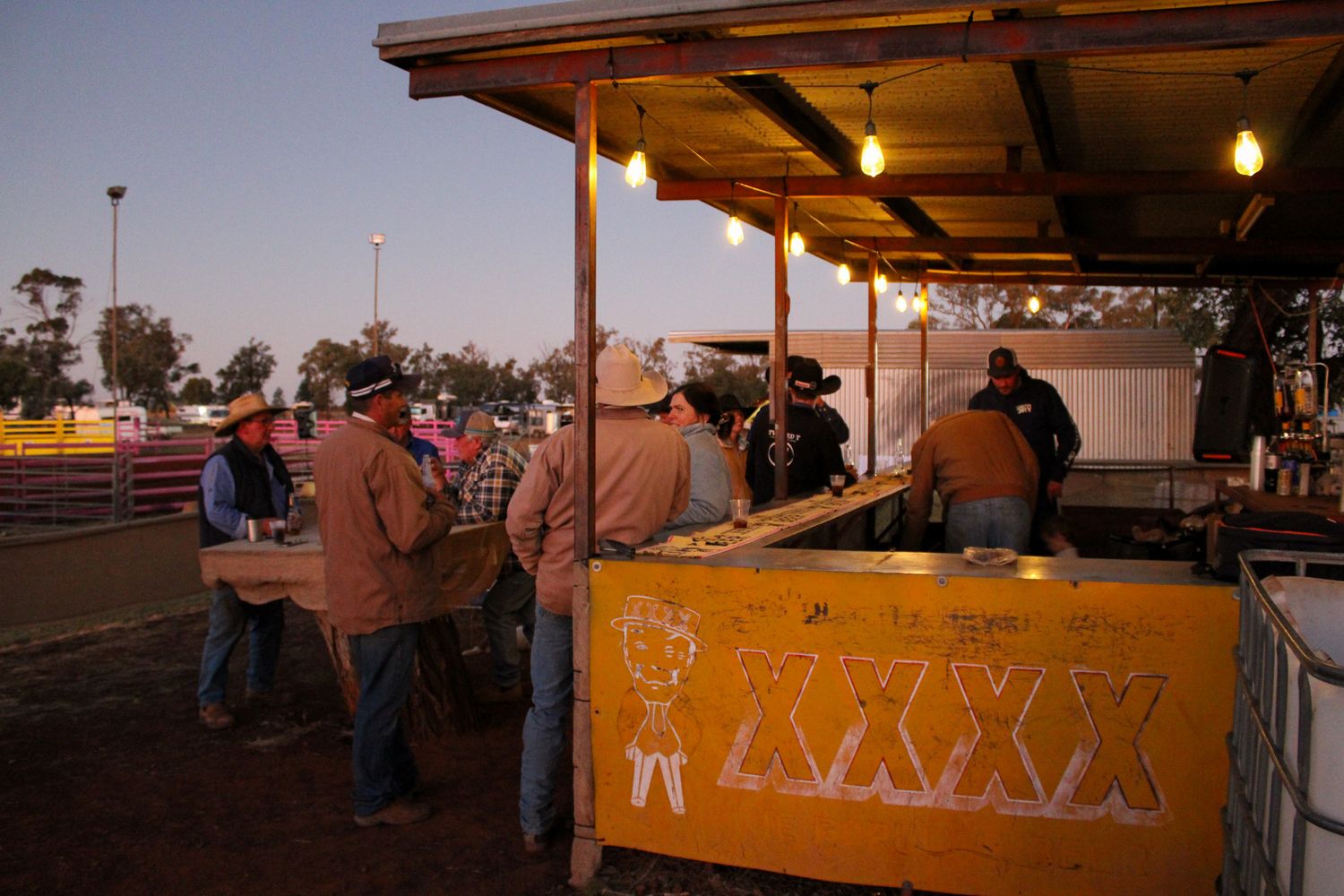 People gather at an outdoor food stand with orange counters and string lights at dusk.
