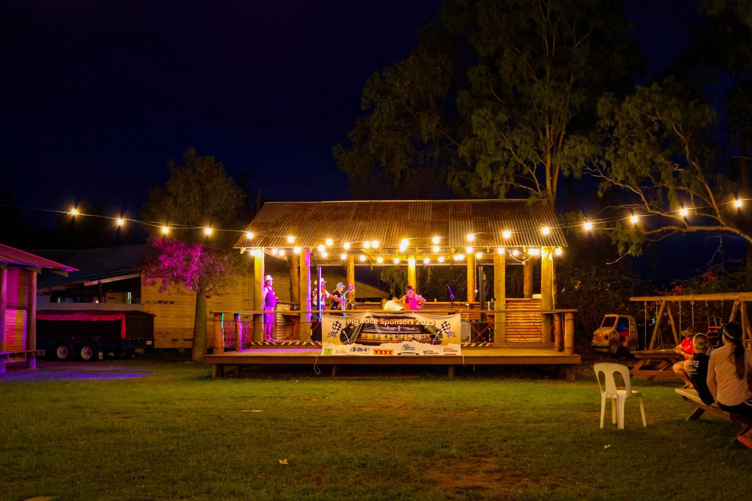 String lights illuminate an outdoor nighttime event space with rustic wooden structures and trees in the background.