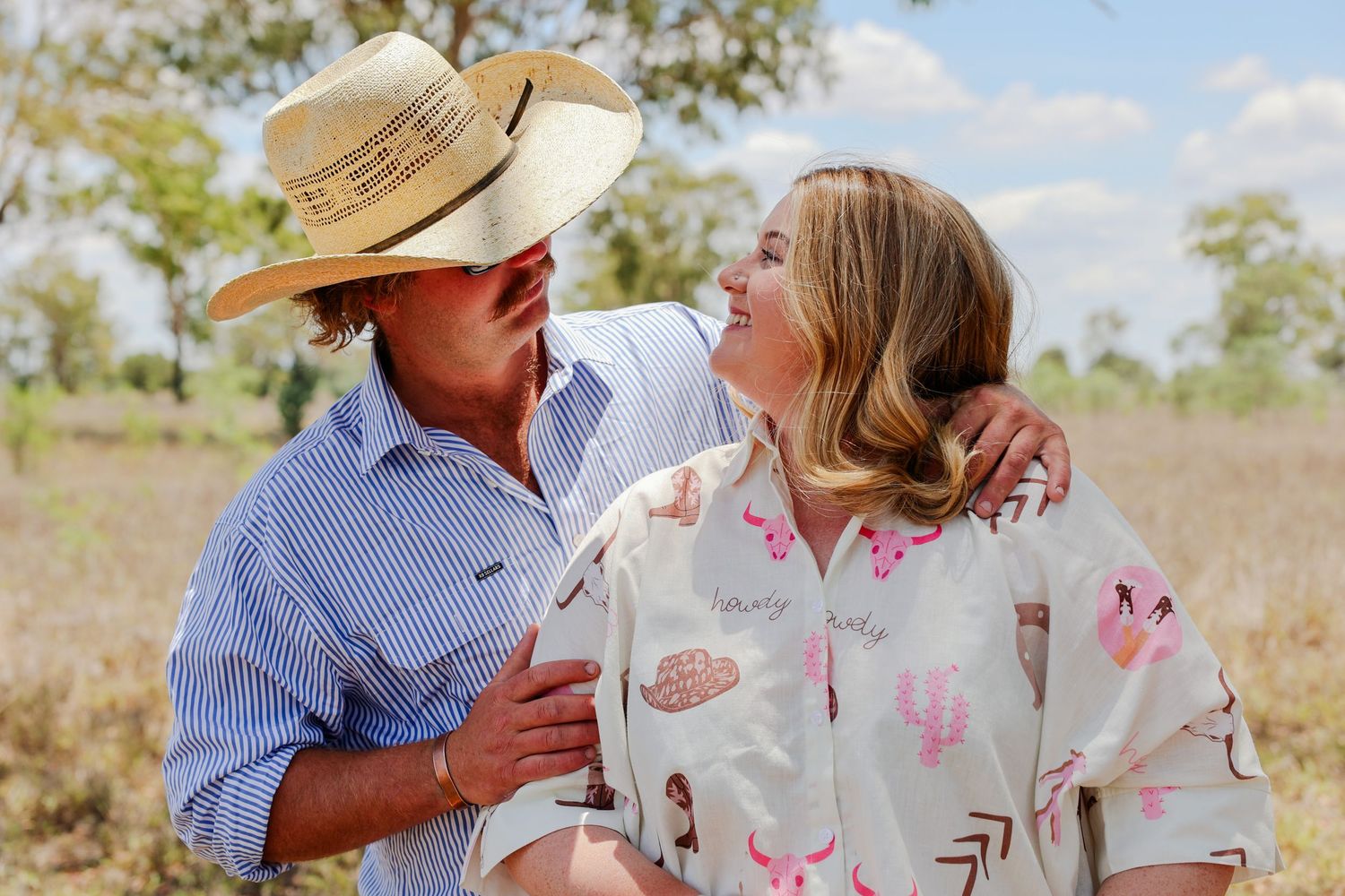 A couple in western attire shares a romantic moment in a sunlit field wearing cowboy hats and casual clothing.