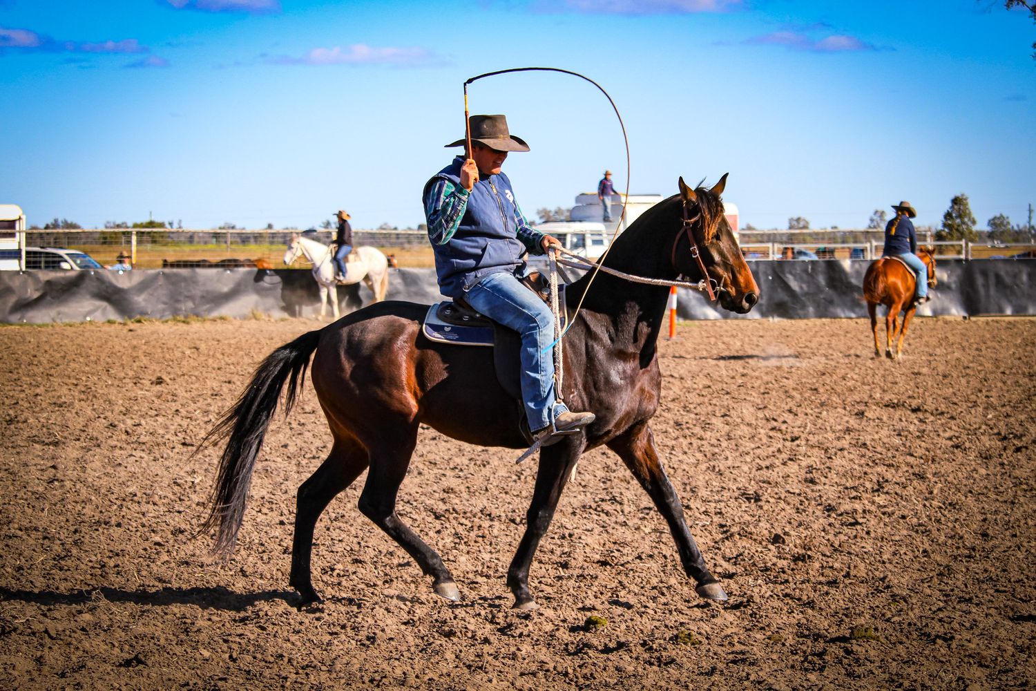 Riders on horseback practicing roping techniques in a dusty arena under blue skies.