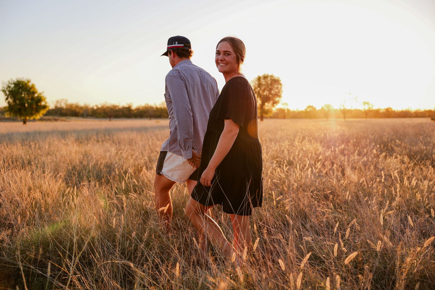 Two people stand together in a golden wheat field at sunset, creating a romantic silhouette against the warm light.