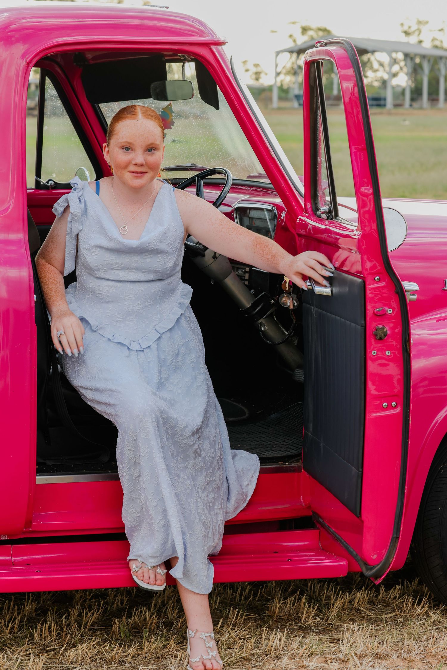 A vintage bright pink pickup truck with the door open showing someone seated in a gray dress.