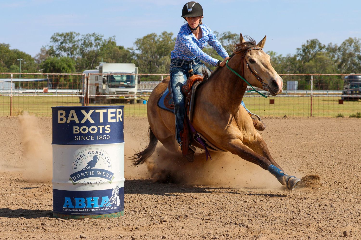 A barrel racing rider on a brown horse rounds a Baxter Boots barrel in a dusty arena during competition.