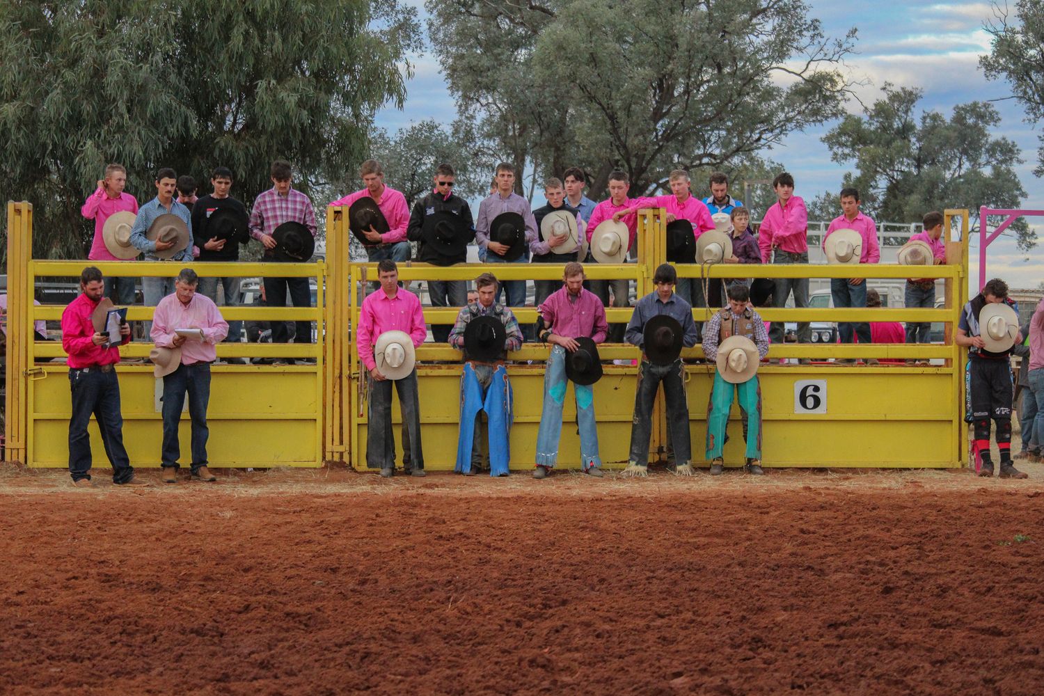 A group of people watching a rodeo event from behind yellow metal barriers on red dirt ground.
