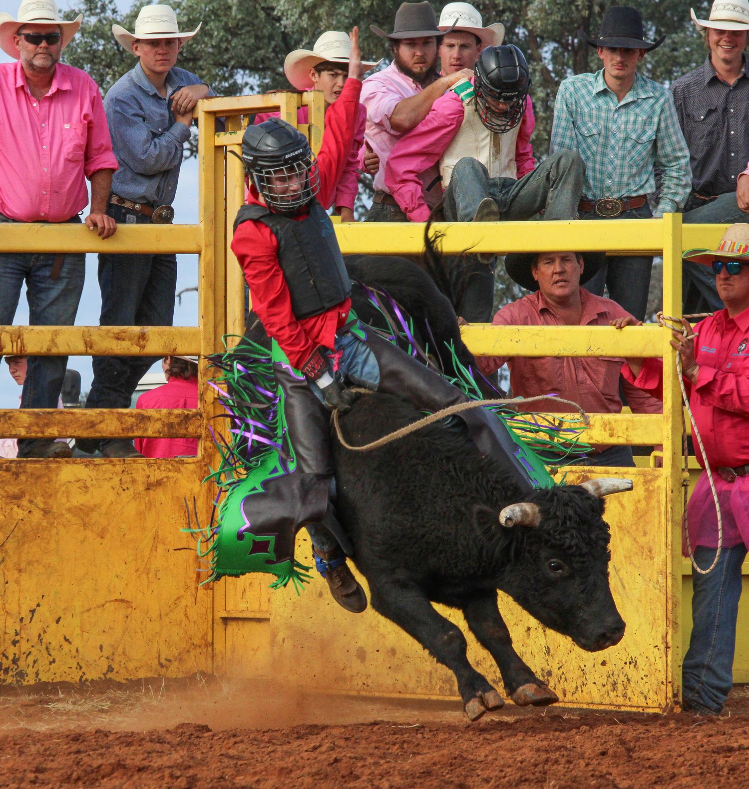 Rodeo bull rider in red and green attire holds on tight as black bull bucks in yellow-fenced arena.