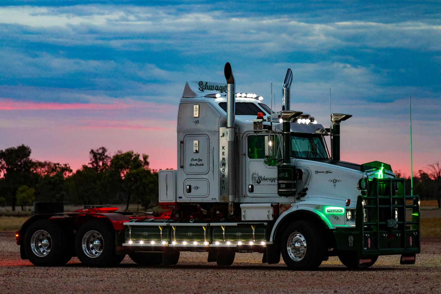 Semi trucks line up against a vibrant sunset sky with colorful clouds and silhouetted trees in the background.