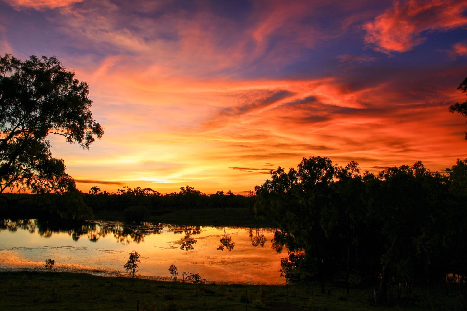 Dramatic orange and purple clouds illuminate the sky during a vibrant sunset over a rural landscape.