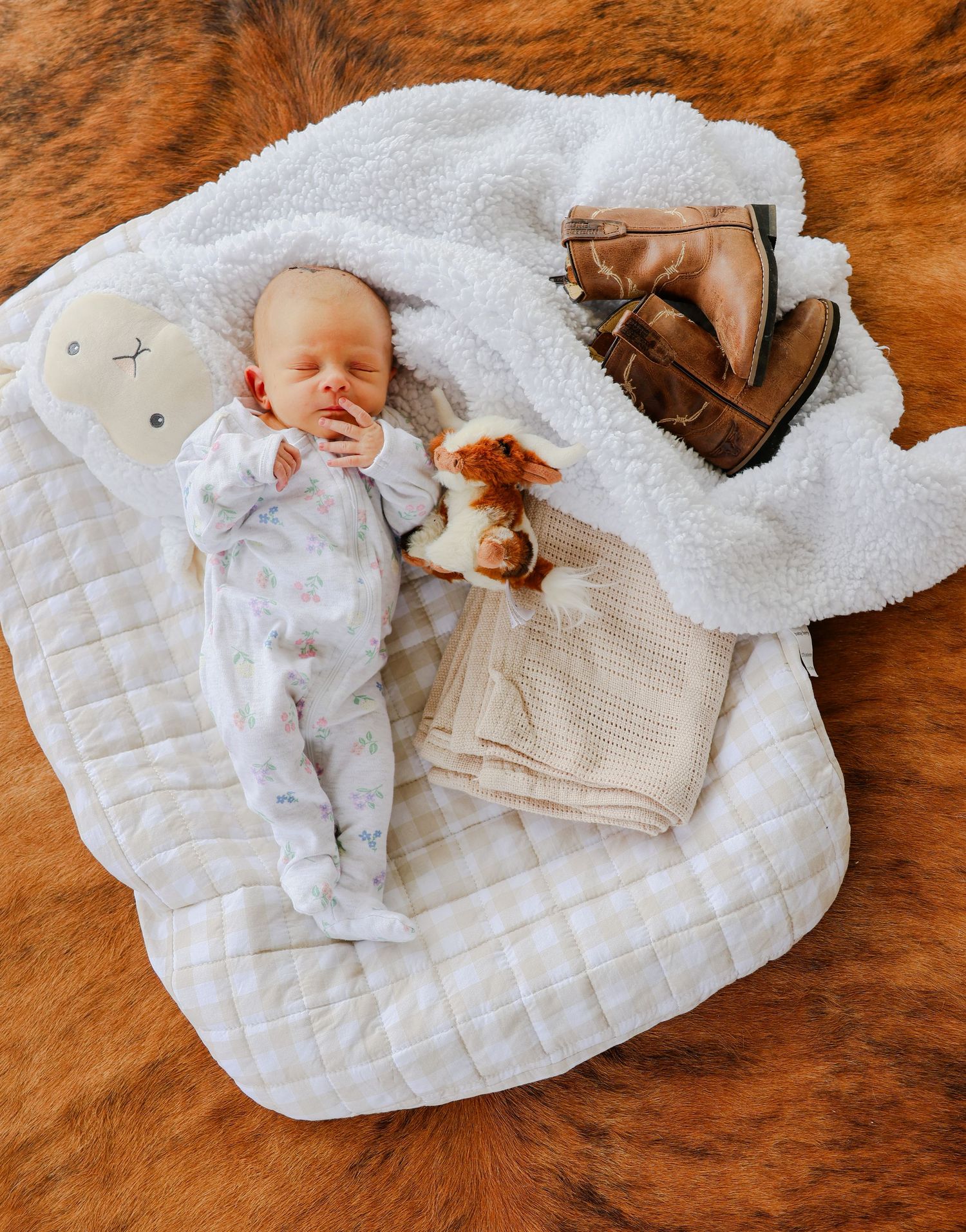 A small baby rests on a white quilted blanket with knit accessories and wooden toys on a wooden surface.