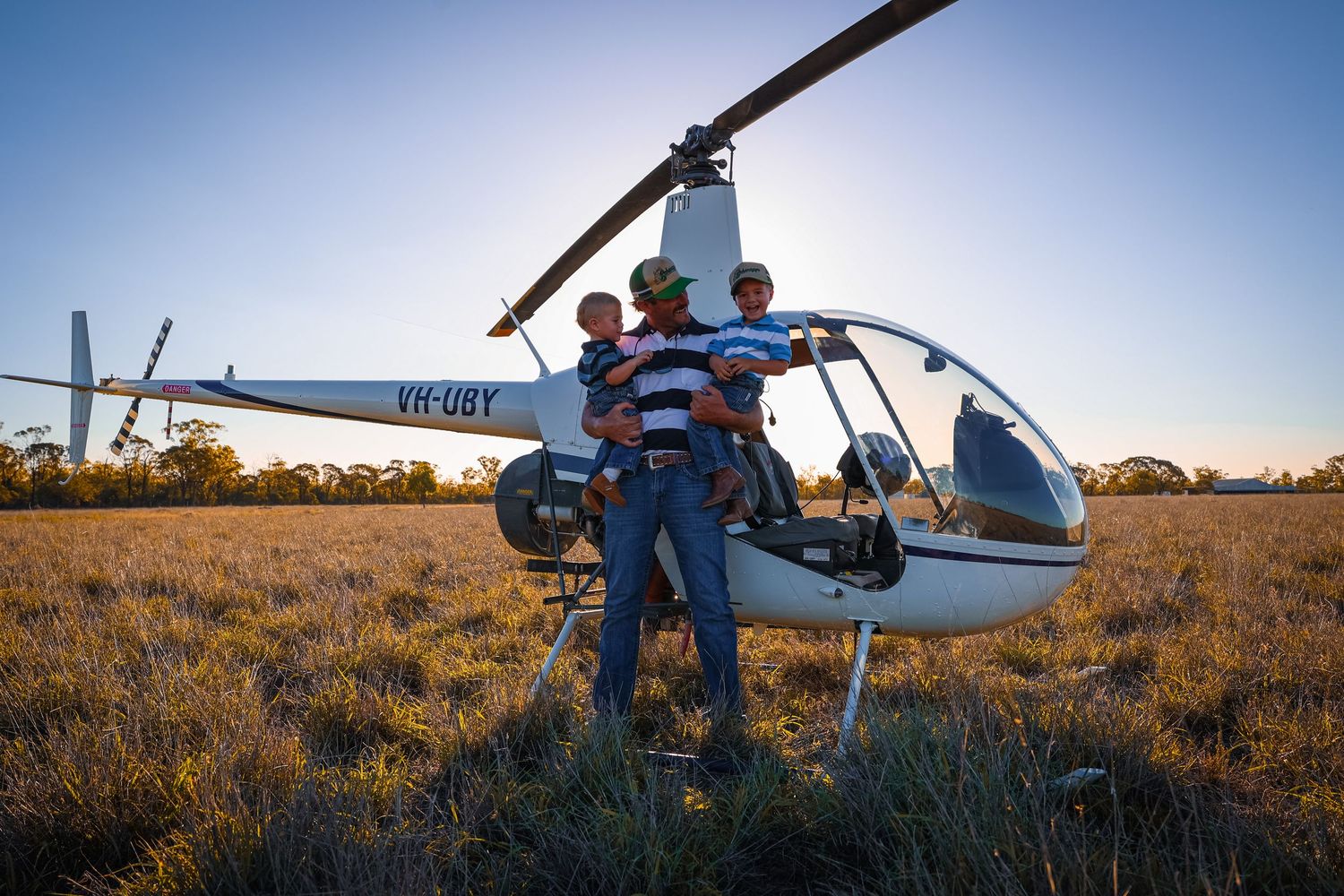 A helicopter lands in a grassy field during sunset while people gather around to observe.