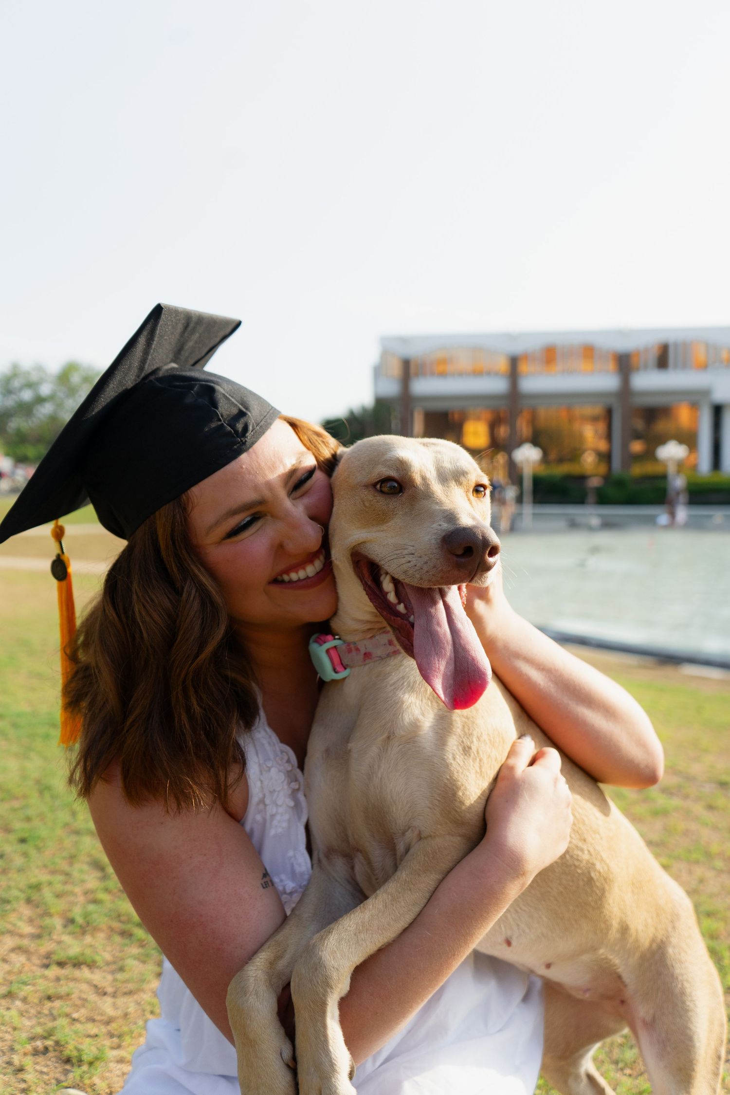 Smiling graduate in cap and white dress hugging a yellow Labrador dog outdoors near a fountain.